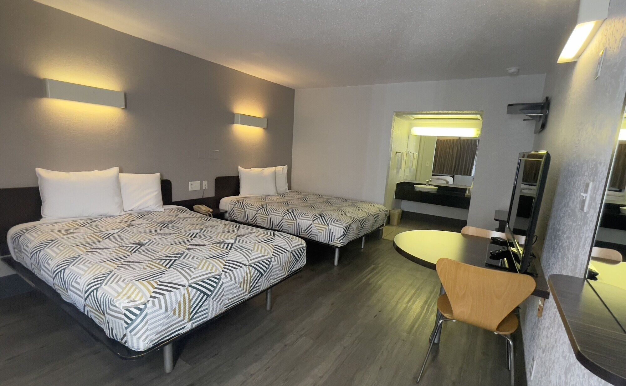A hotel room with two beds, a small desk, a chair, a round table, and a bathroom visible through an open door.