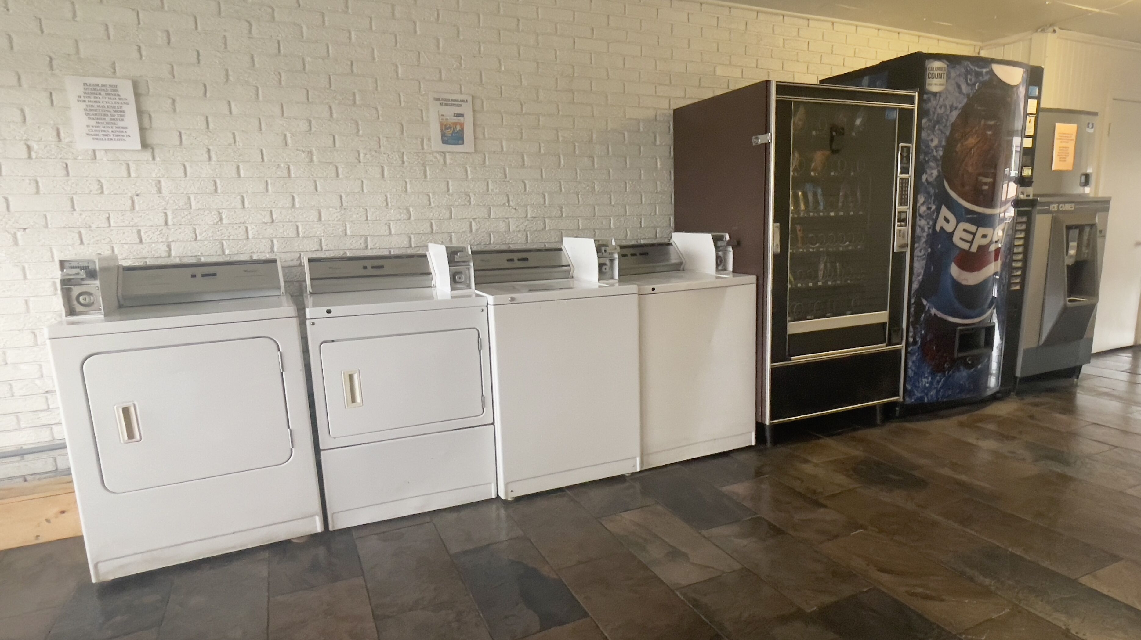 A row of white laundry machines in a public area with a Pepsi vending machine to the right.