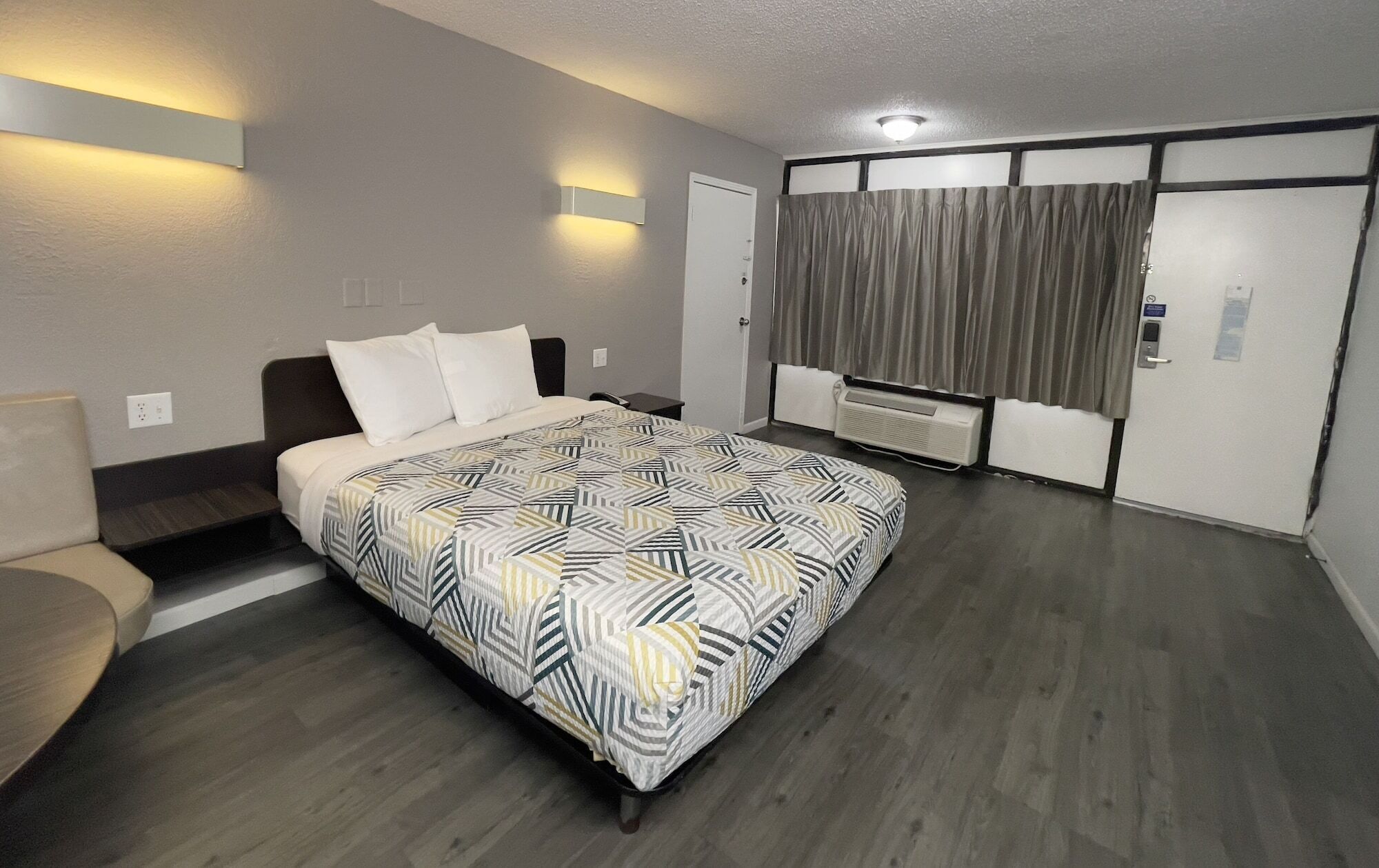 A hotel room with a bed, a bedside table, a chair, a window with curtains, a wall-mounted air conditioner, and a door.