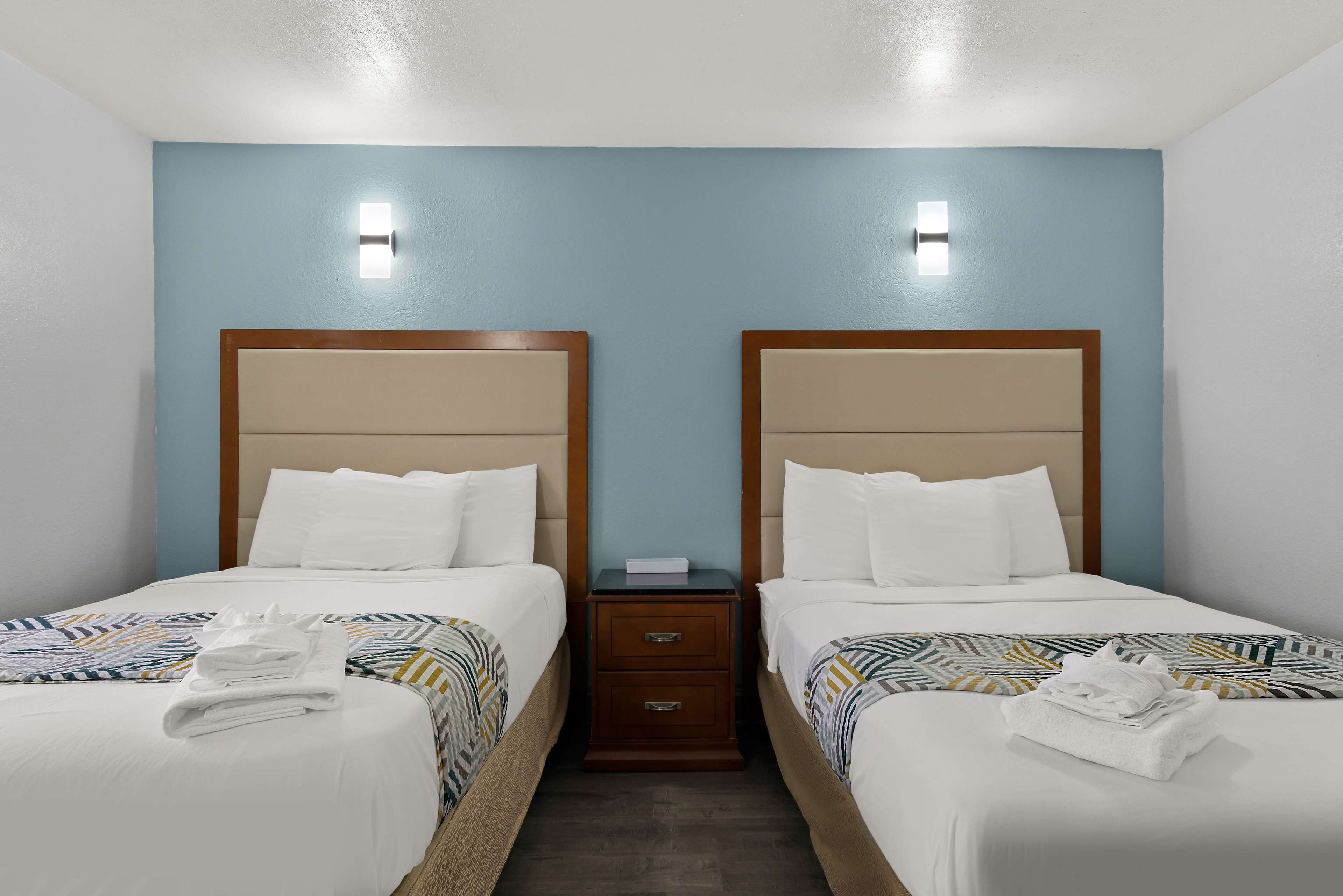 Quarto de hotel com duas camas, cada uma com cabeceira de madeira, roupa de cama branca e manta estampada. Há duas mesas de cabeceira com abajures e uma pequena gaveta.