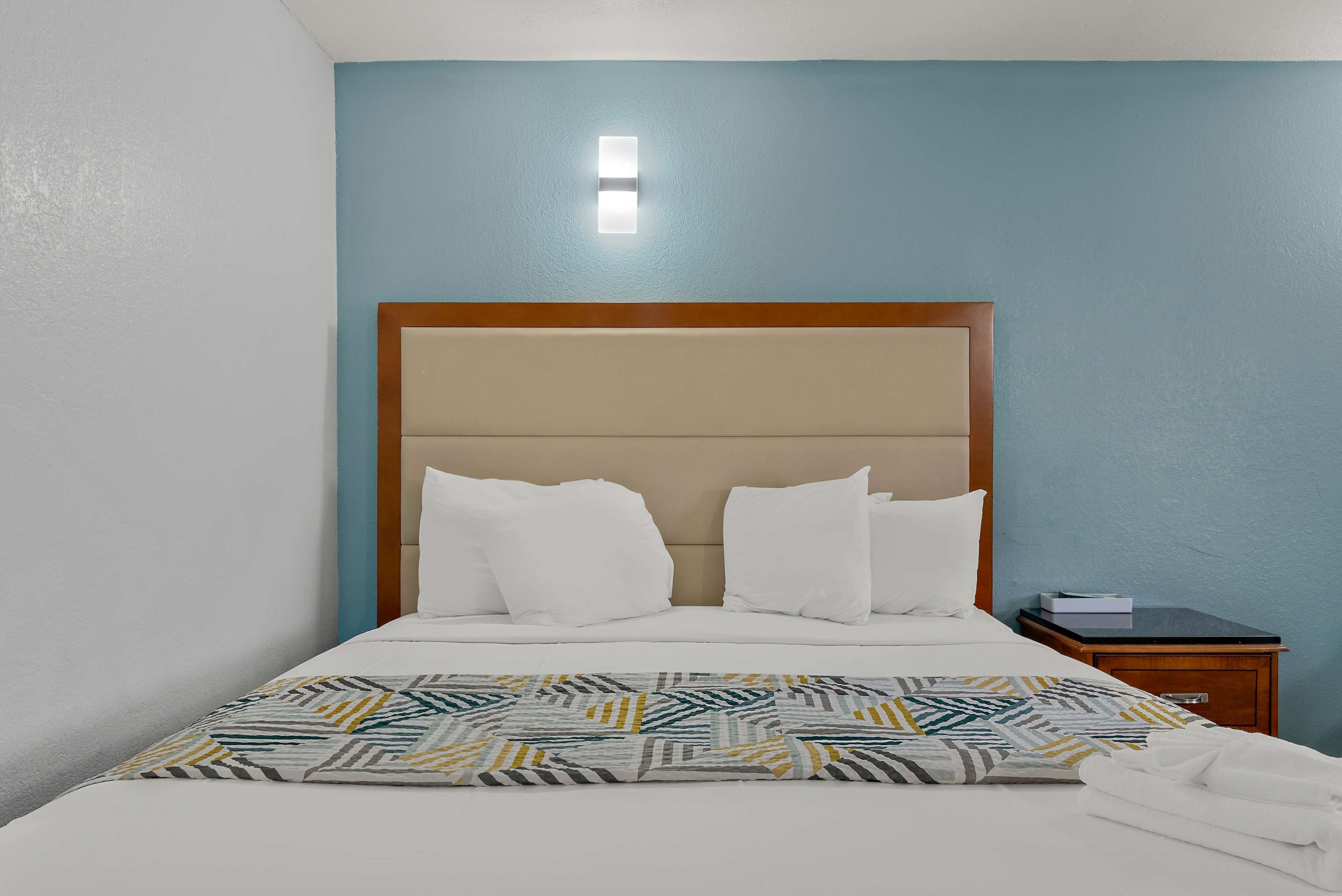 Una cama bien hecha con sábanas blancas y un plaid estampado, una mesita de noche de madera con un libro, y una lámpara de pared.