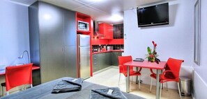 A compact room with a kitchenette, a dining area with red chairs, a TV mounted on the wall, and a bed with a gray blanket.