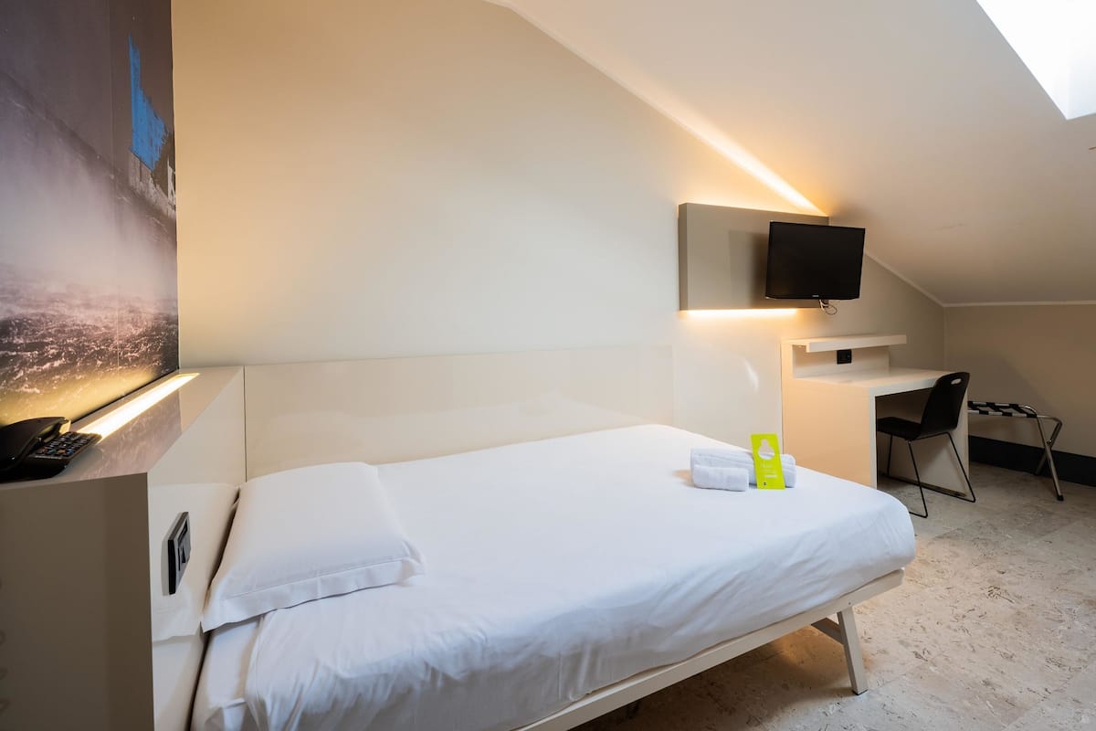 A hotel room with a bed, a desk with a chair, a TV mounted on the wall, and a painting on the wall.