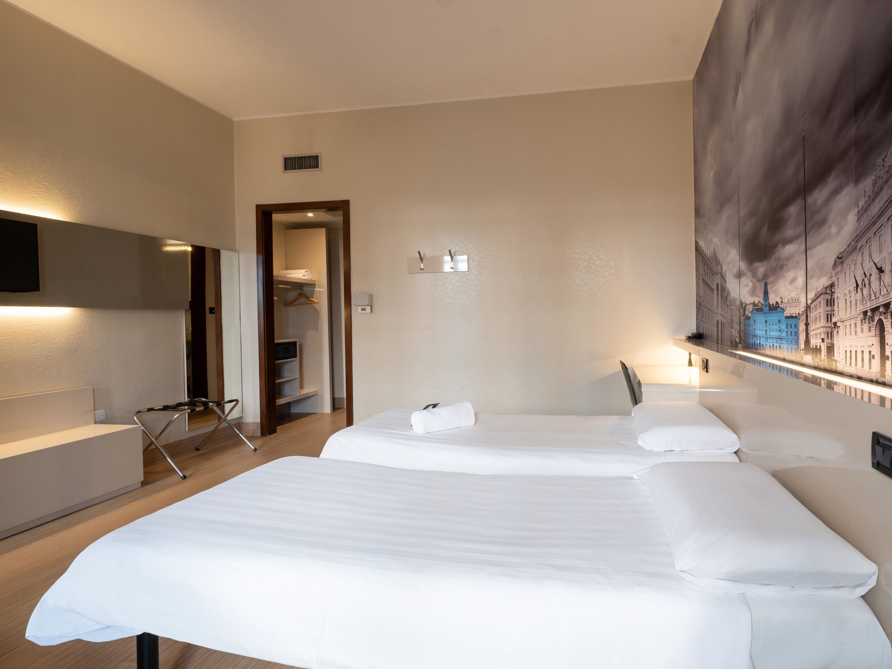 A hotel room with two beds, a walk-in closet, a desk, and a large cityscape mural on the wall.