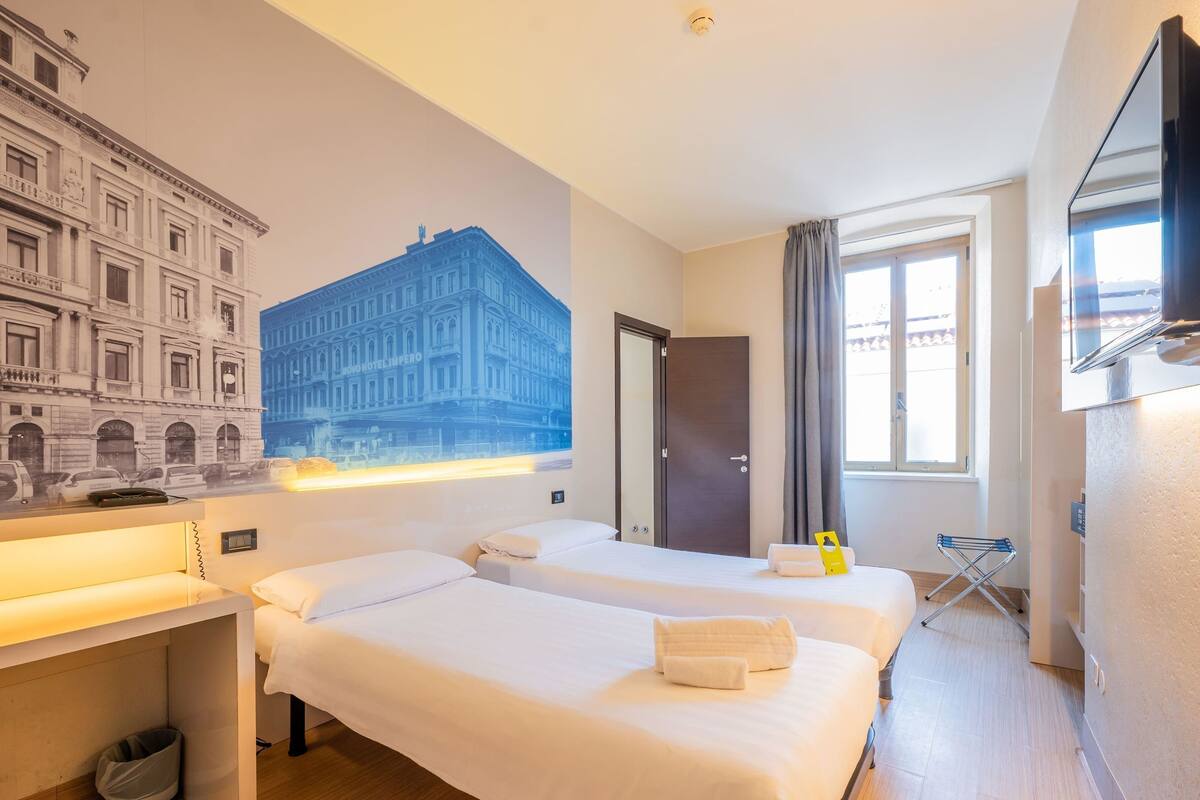 A hotel room with two beds, a desk, a chair, a TV, a window with curtains, and a wall mural.