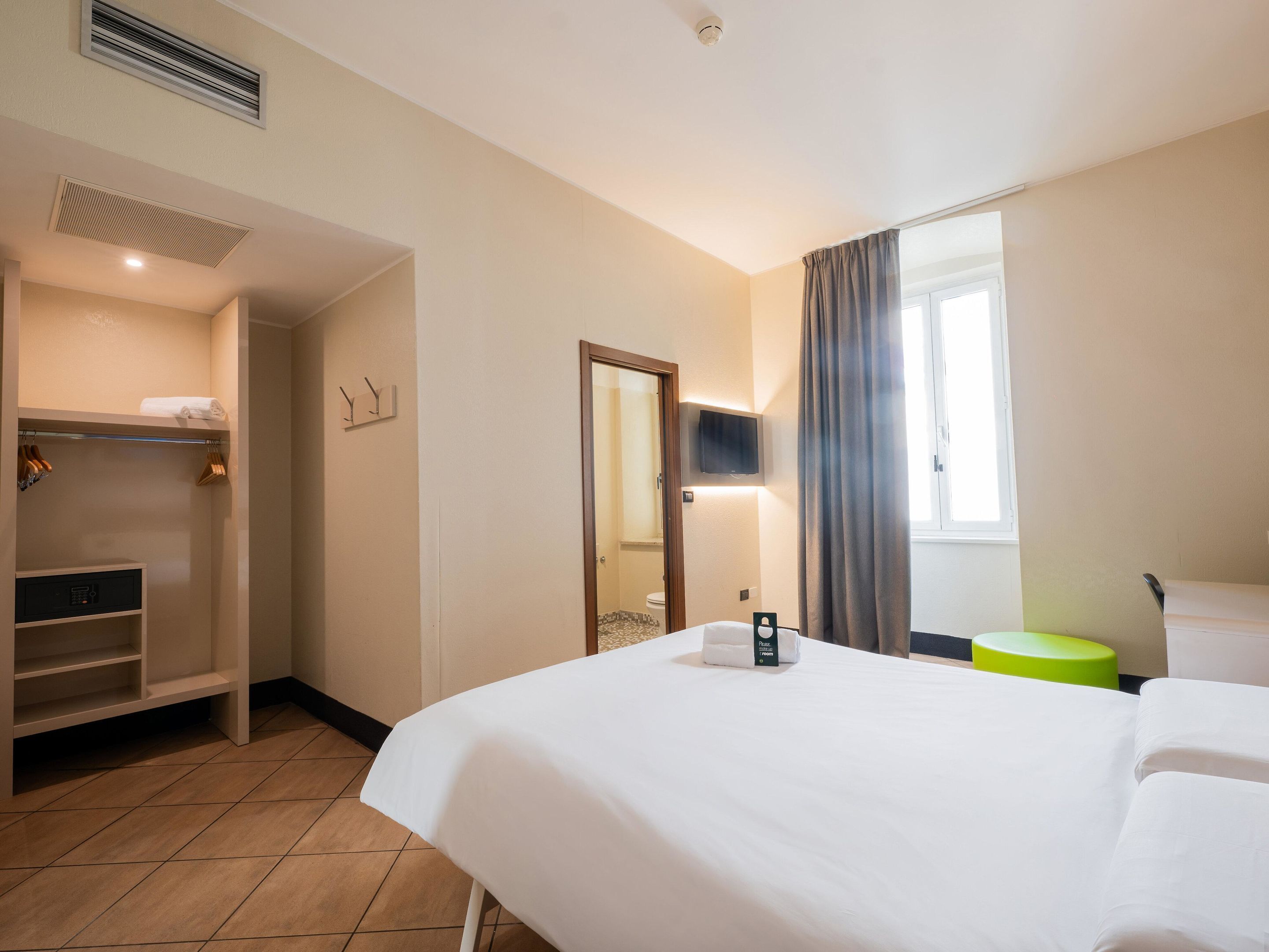 A hotel room with a bed, a wardrobe, a mirror, a television, and a window with curtains.