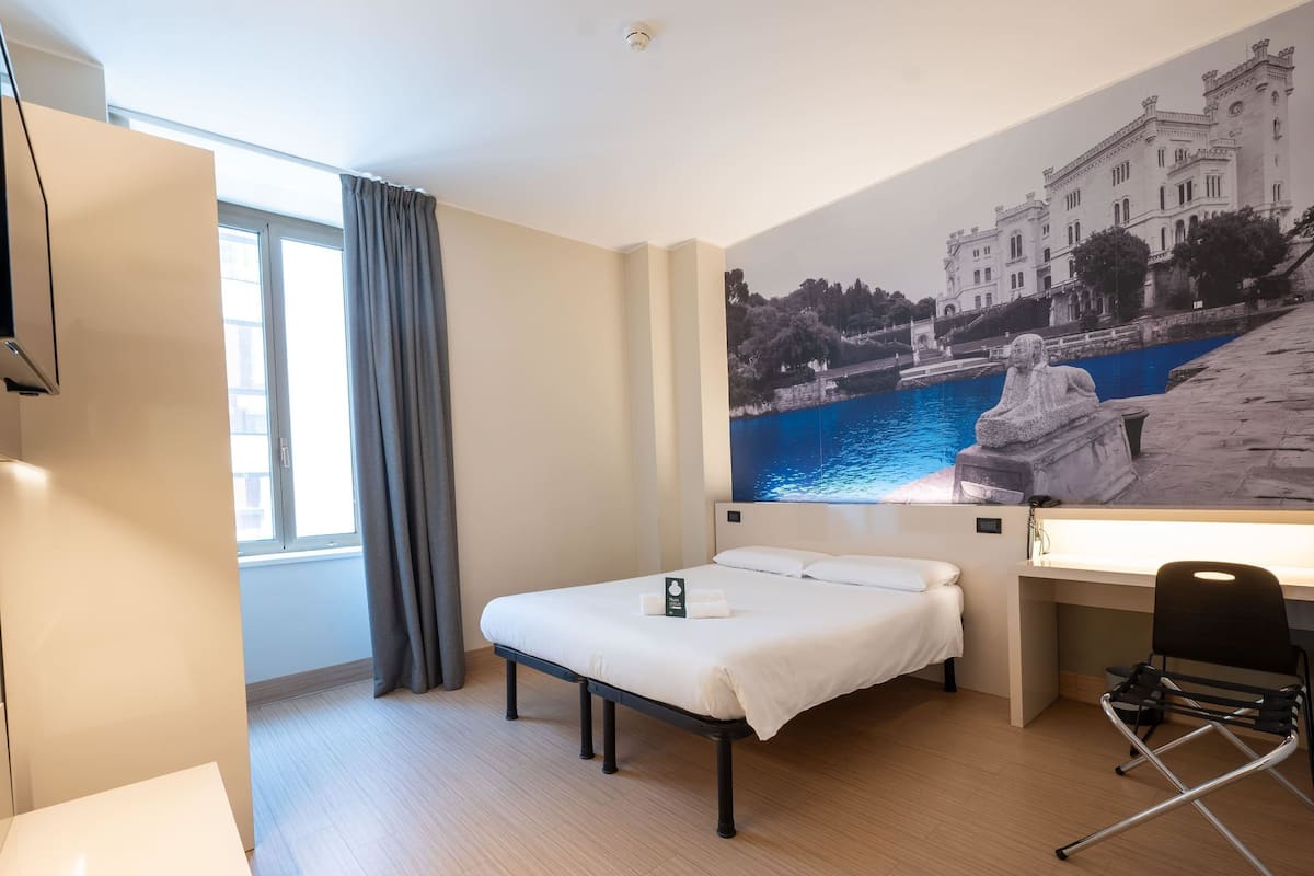 A hotel room with a bed, a desk, a chair, a window with curtains, and a large wall mural of a building by a body of water.