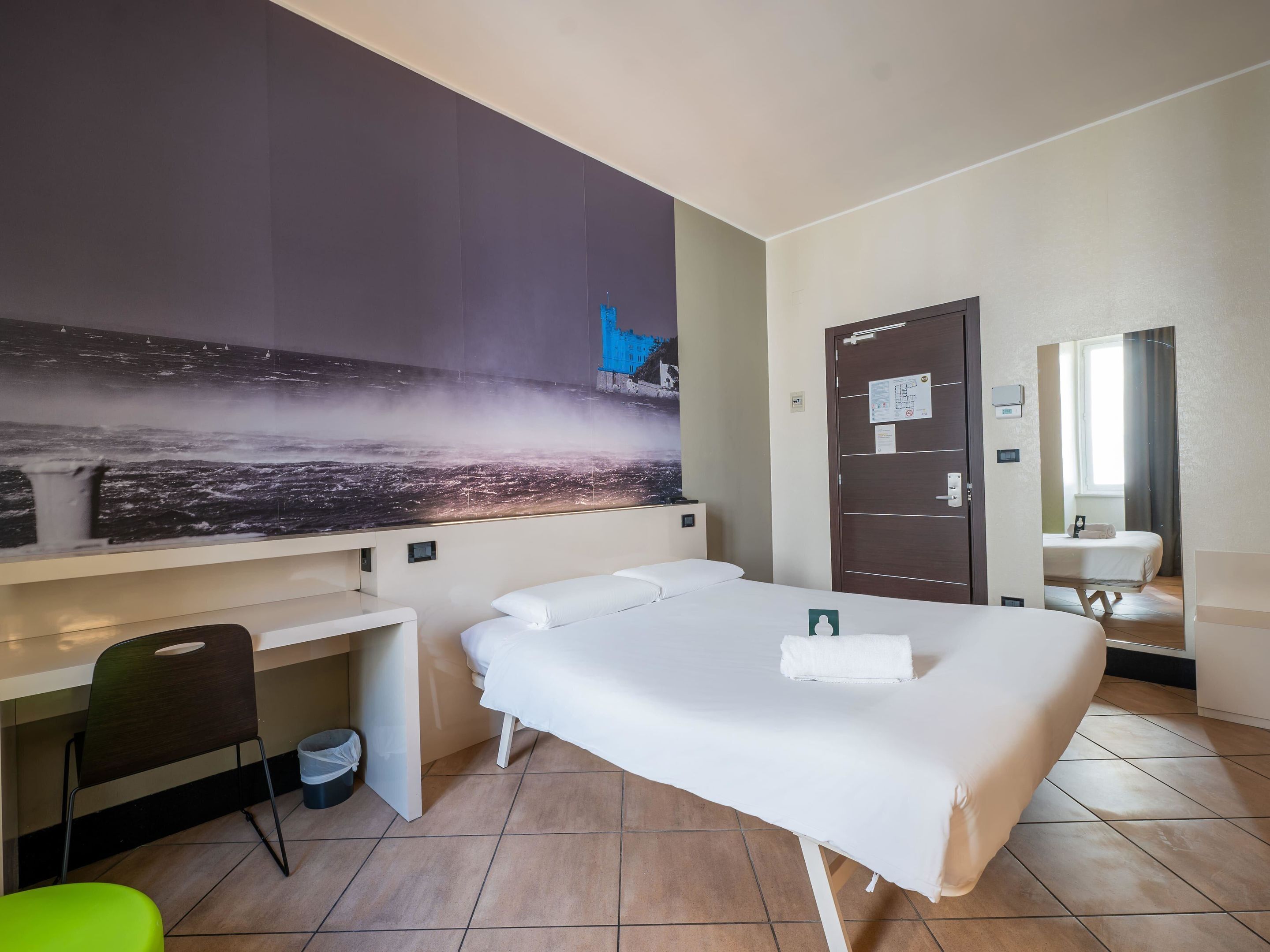 A hotel room with a bed, a desk, a chair, a large window, and a bathroom mirror.
