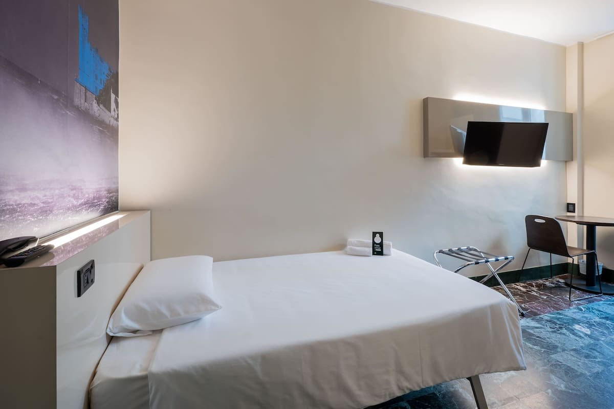A hotel room with a bed, a desk, a chair, a flat-screen TV, and a painting on the wall.