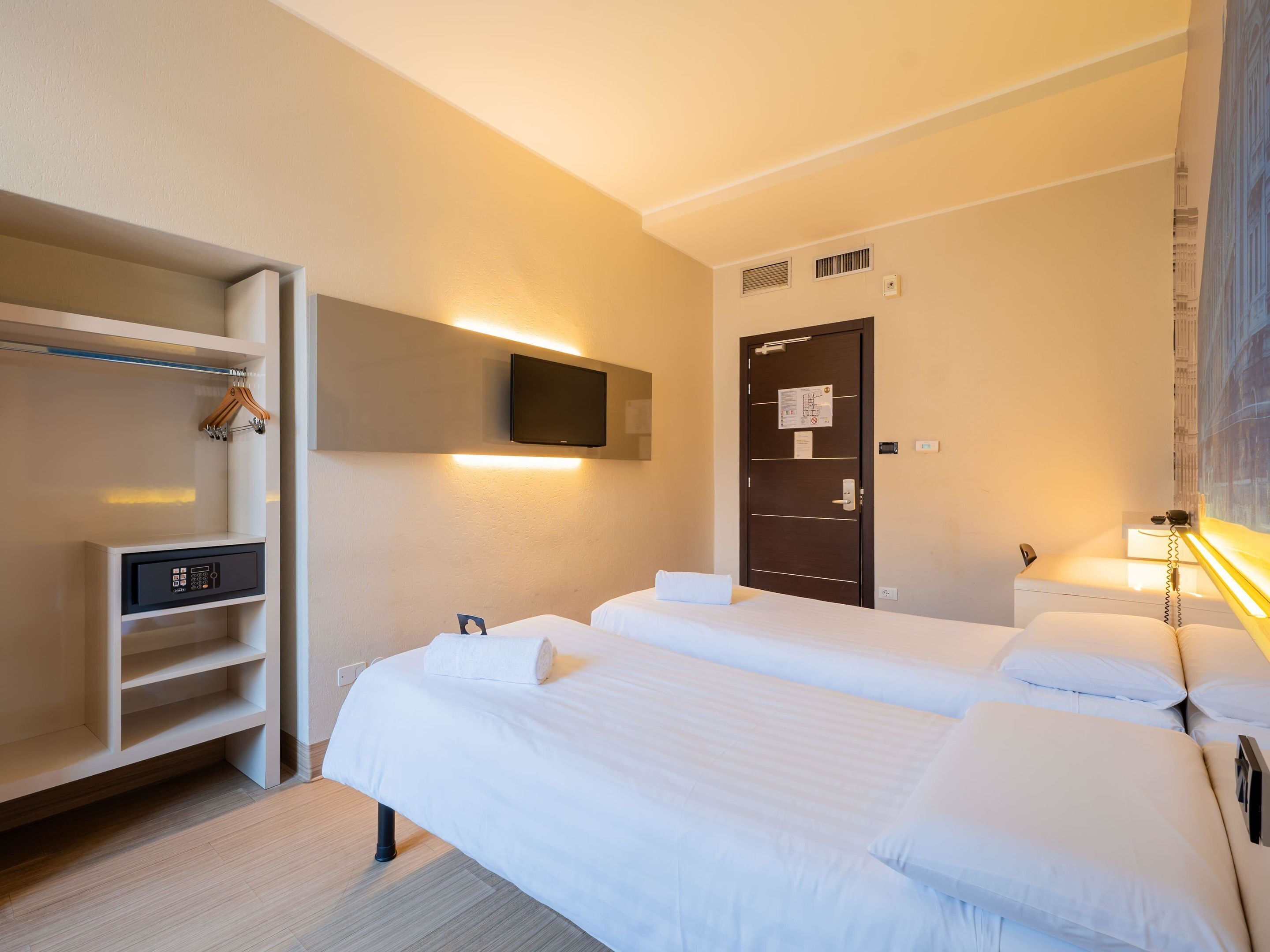 A hotel room with two beds, a wardrobe, a TV, and a desk with a control panel.
