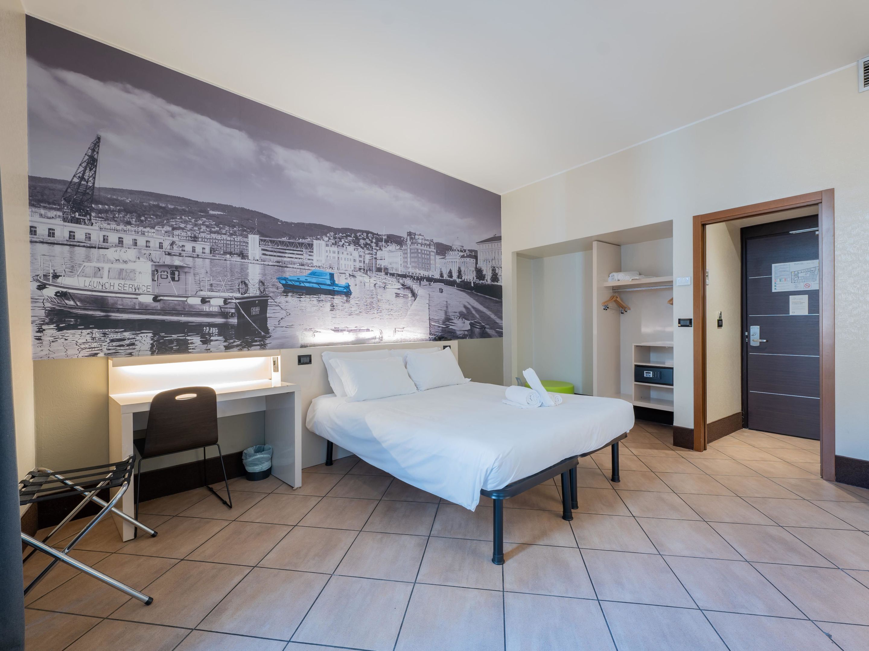 Hotelkamer met een bed, een bureau, een stoel, een kledingkast en een groot stadsgezicht op de muur.