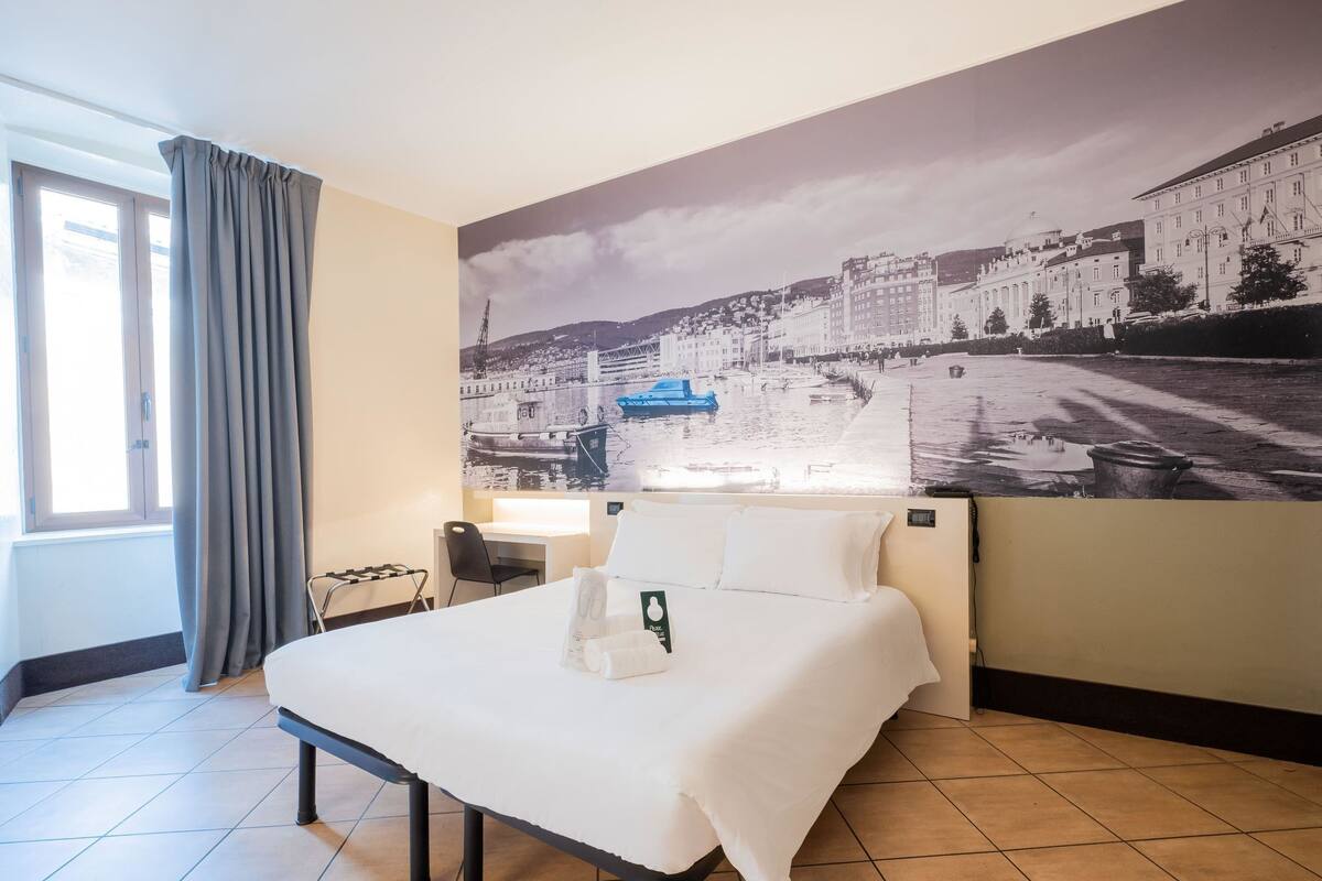 A hotel room with a bed, a desk, a chair, and a large cityscape mural on the wall.