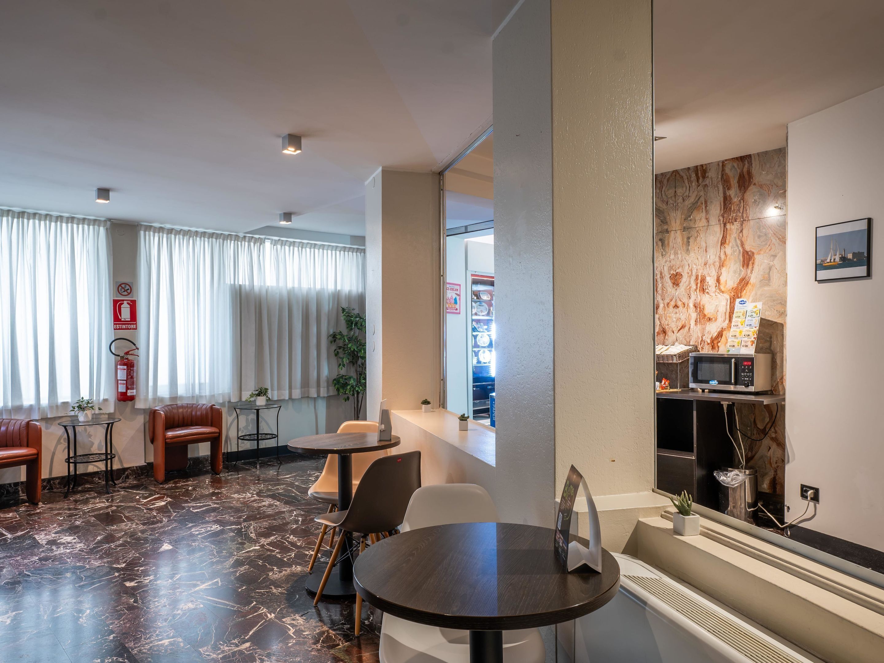 Hall d'albergo con pavimento in marmo, sedute in pelle, un piccolo tavolo con sedie, un forno a microonde, una macchina distributrice e un estintore.