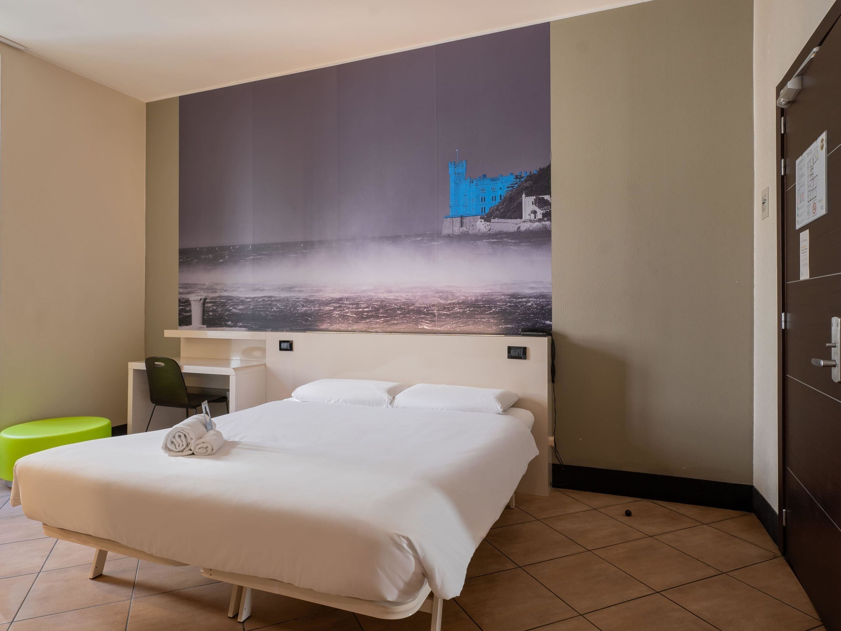 Een hotelkamer met een bed, een bureau en een stoel. Op de muur hangt een groot schilderij waarop een kasteel en een kasteelmuur afgebeeld zijn.