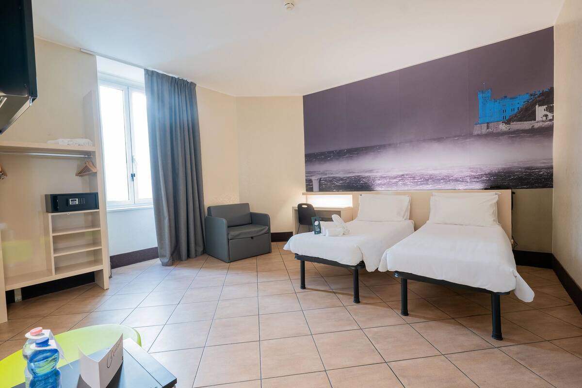 A hotel room with two beds, a desk with a chair, a wardrobe, and a large window with curtains.