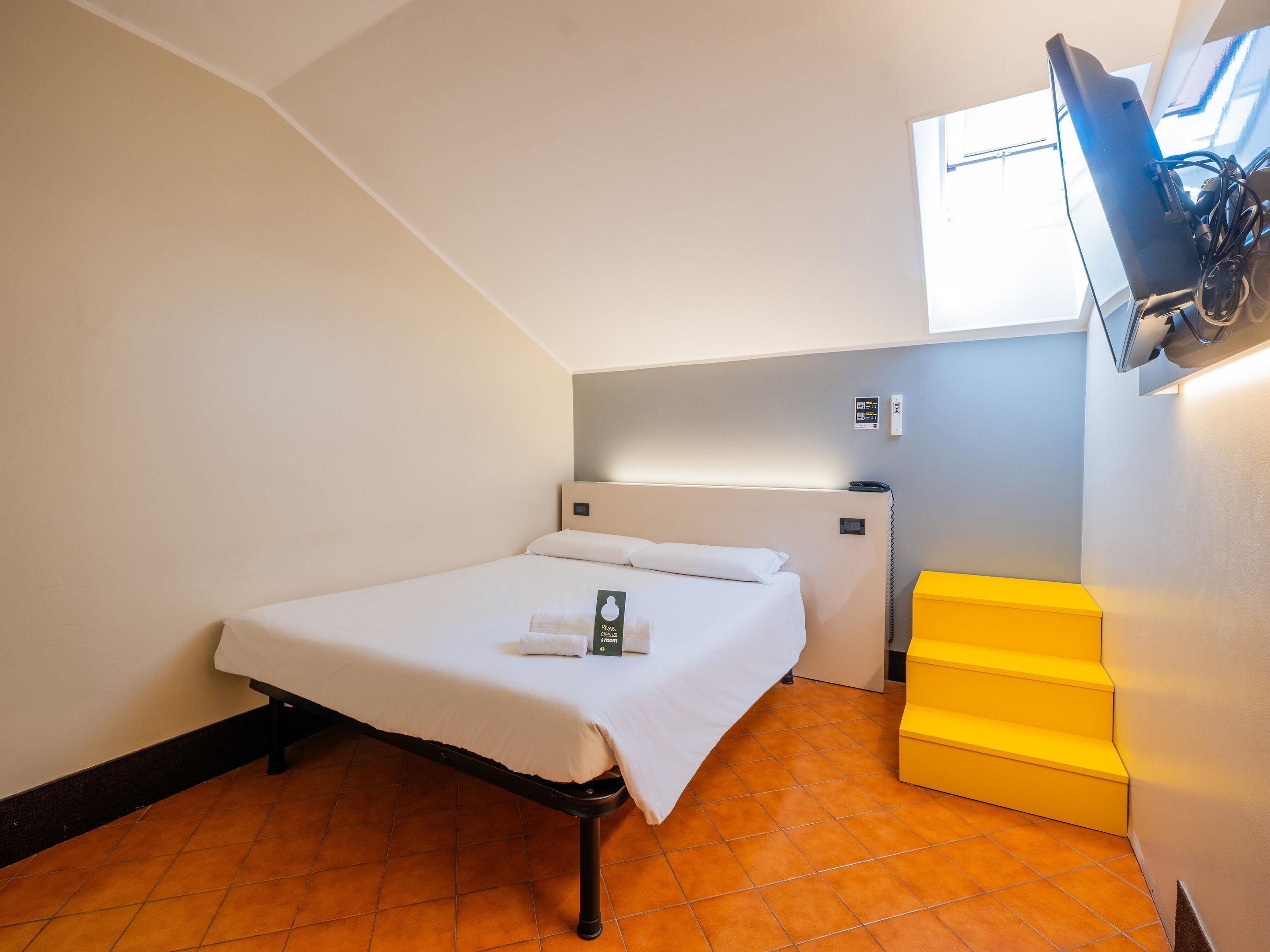A hotel room with a bed, a TV mounted on the wall, a small desk, and a yellow storage box.