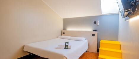A hotel room with a bed, a TV mounted on the wall, a small desk, and a yellow storage box.