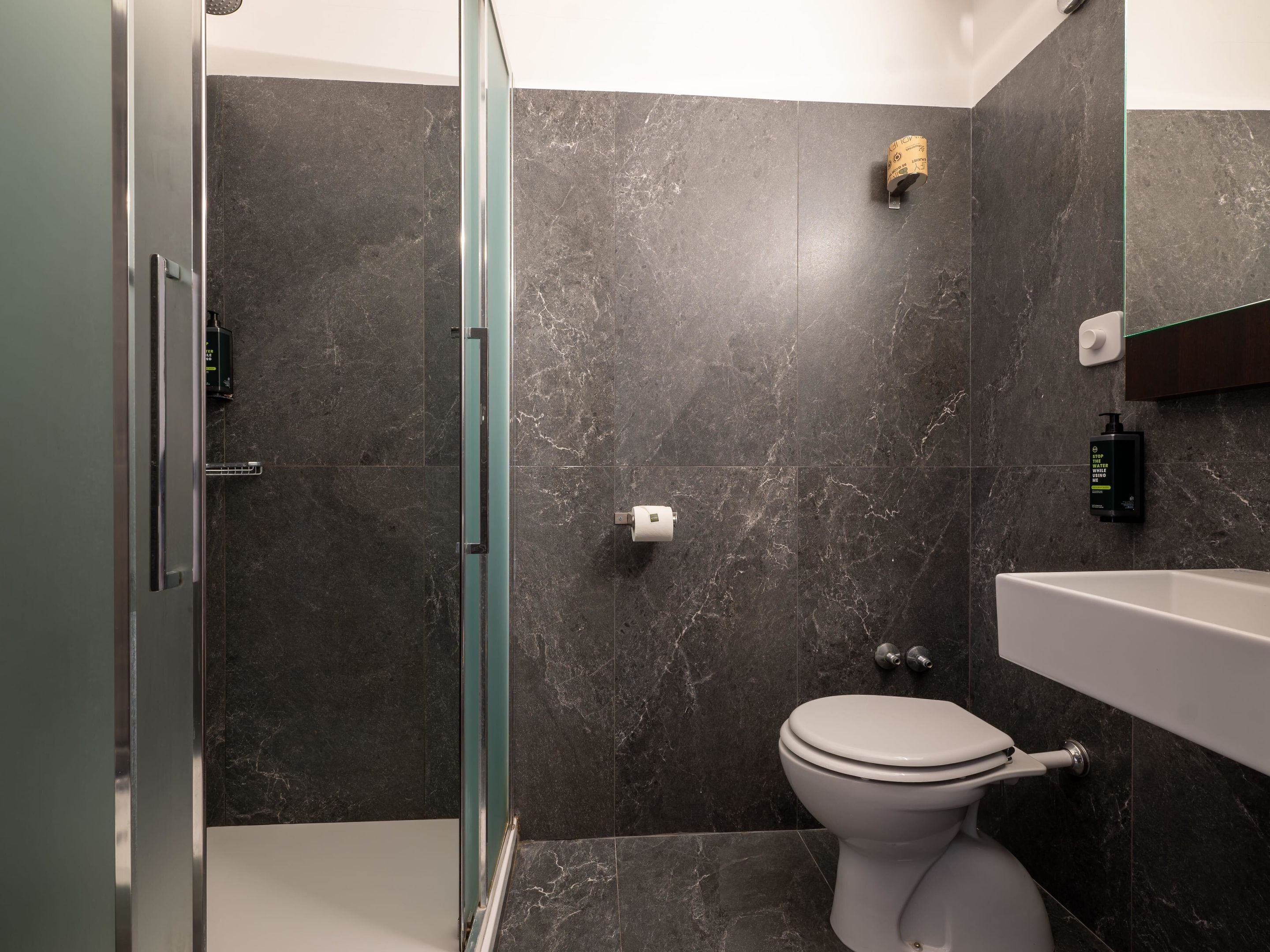 A modern bathroom with dark marble tiles, a glass shower enclosure, a white toilet, and a built-in sink.