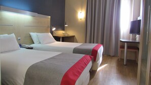 A hotel room with two beds, a desk, a TV, and a window with curtains.