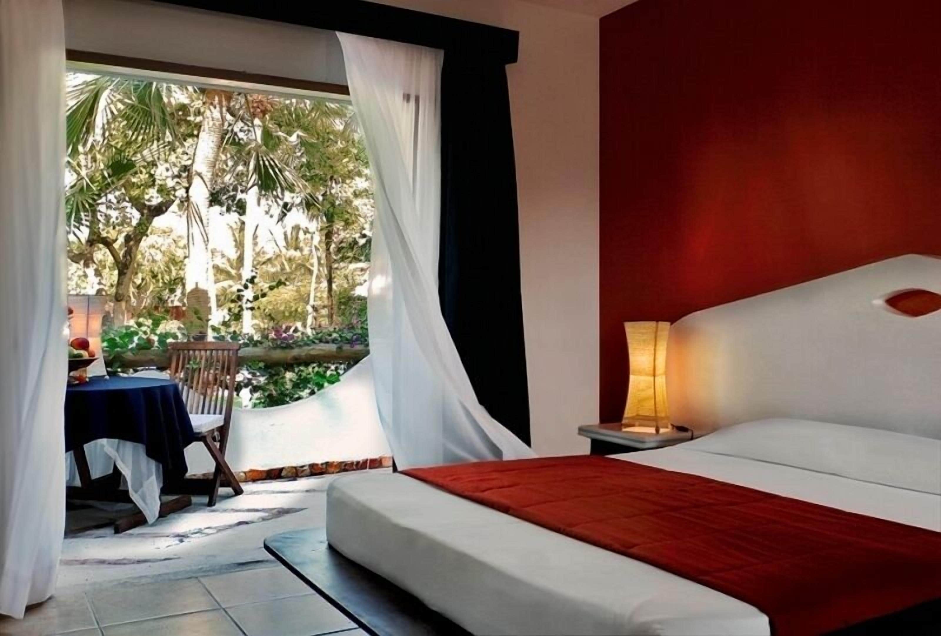 A bedroom with a bed, a nightstand, a table with chairs, and a view of palm trees through sheer curtains.