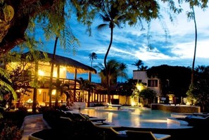A poolside area with lounge chairs, a covered seating area, and a building with warm lighting.