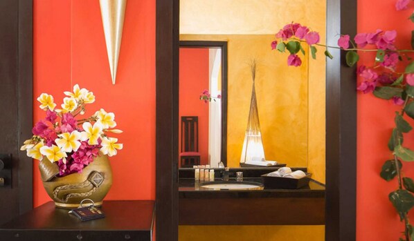 A room with a vibrant orange wall, a decorative vase with flowers, a mirror reflecting a tall lamp, and a small table with a book and a phone. - My Blue Hotel (Jijoca de Jericoacoara)