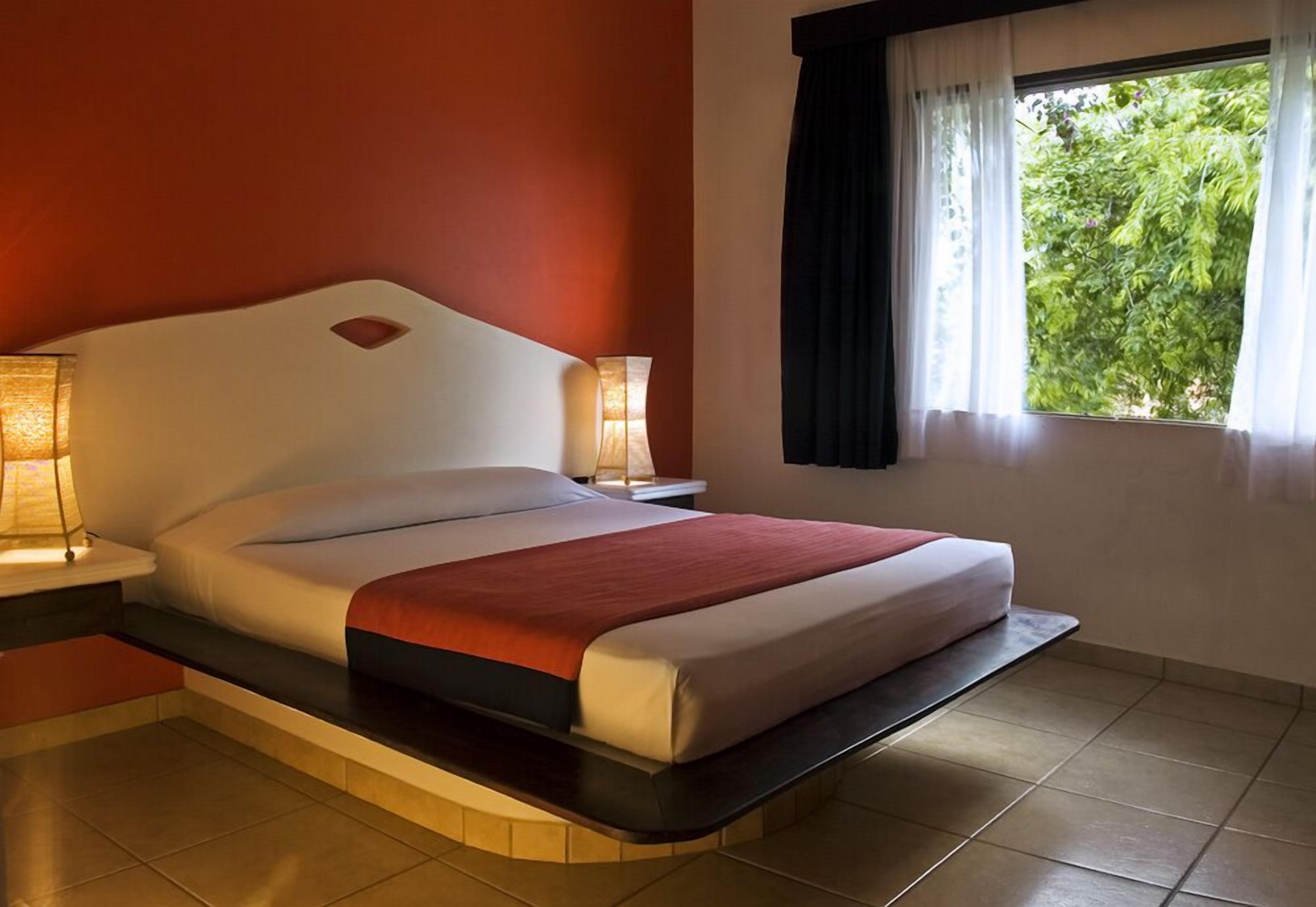 Habitación de hotel moderna con cama curvada, dos lámparas de noche, ventana con cortinas y vista a zonas verdes.