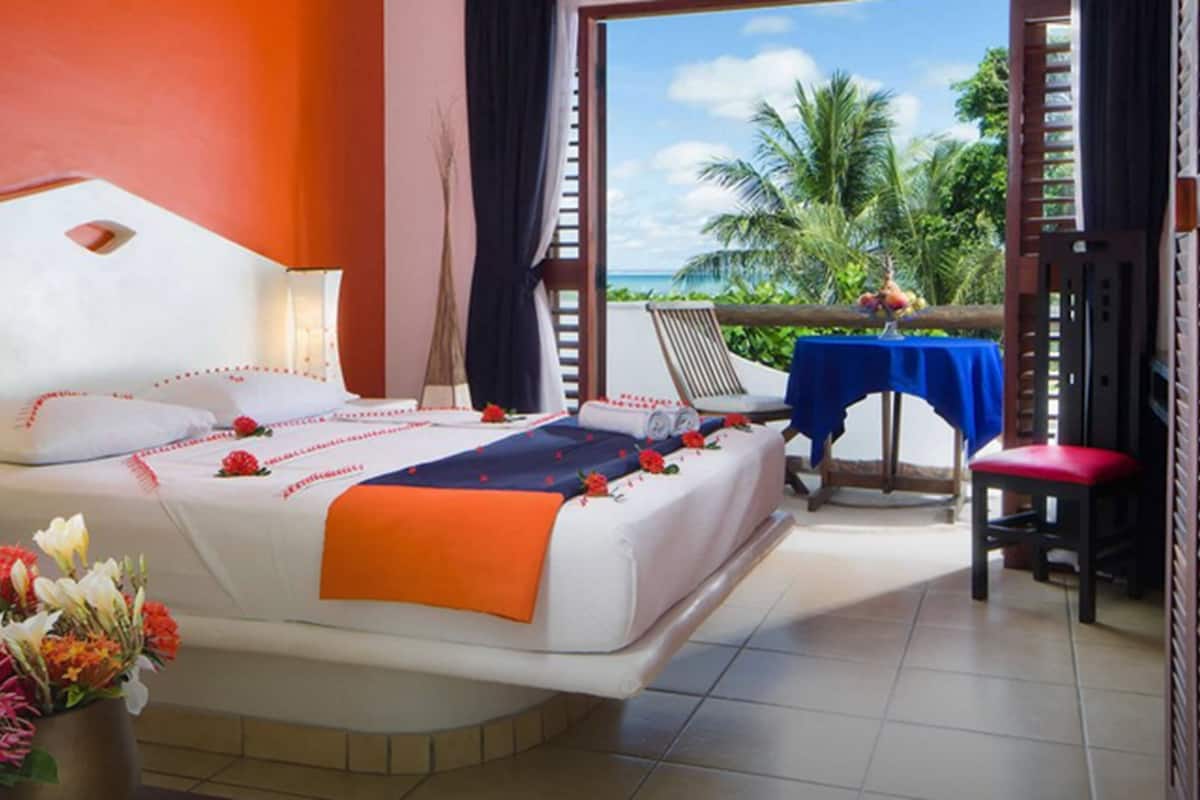 A hotel room with a bed, a chair, a table with a blue tablecloth, and a view of the ocean and palm trees.