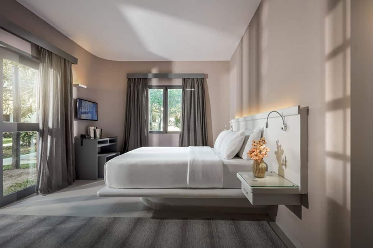 A modern hotel room with a large bed, a bedside table with a vase, a TV mounted on the wall, and a window with curtains.