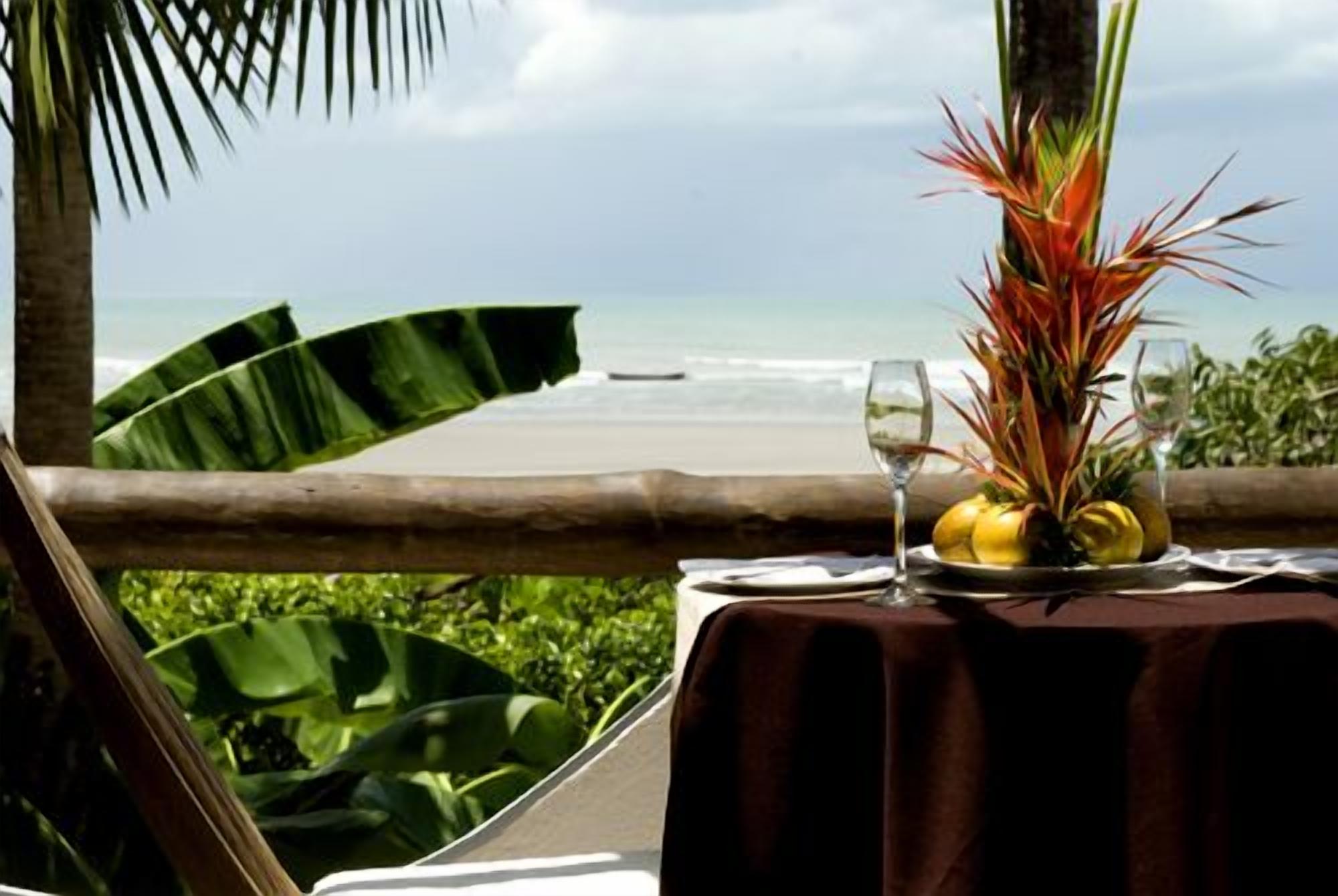 Una mesa con una composición de frutas tropicales y un vaso de agua, con vista a una playa.