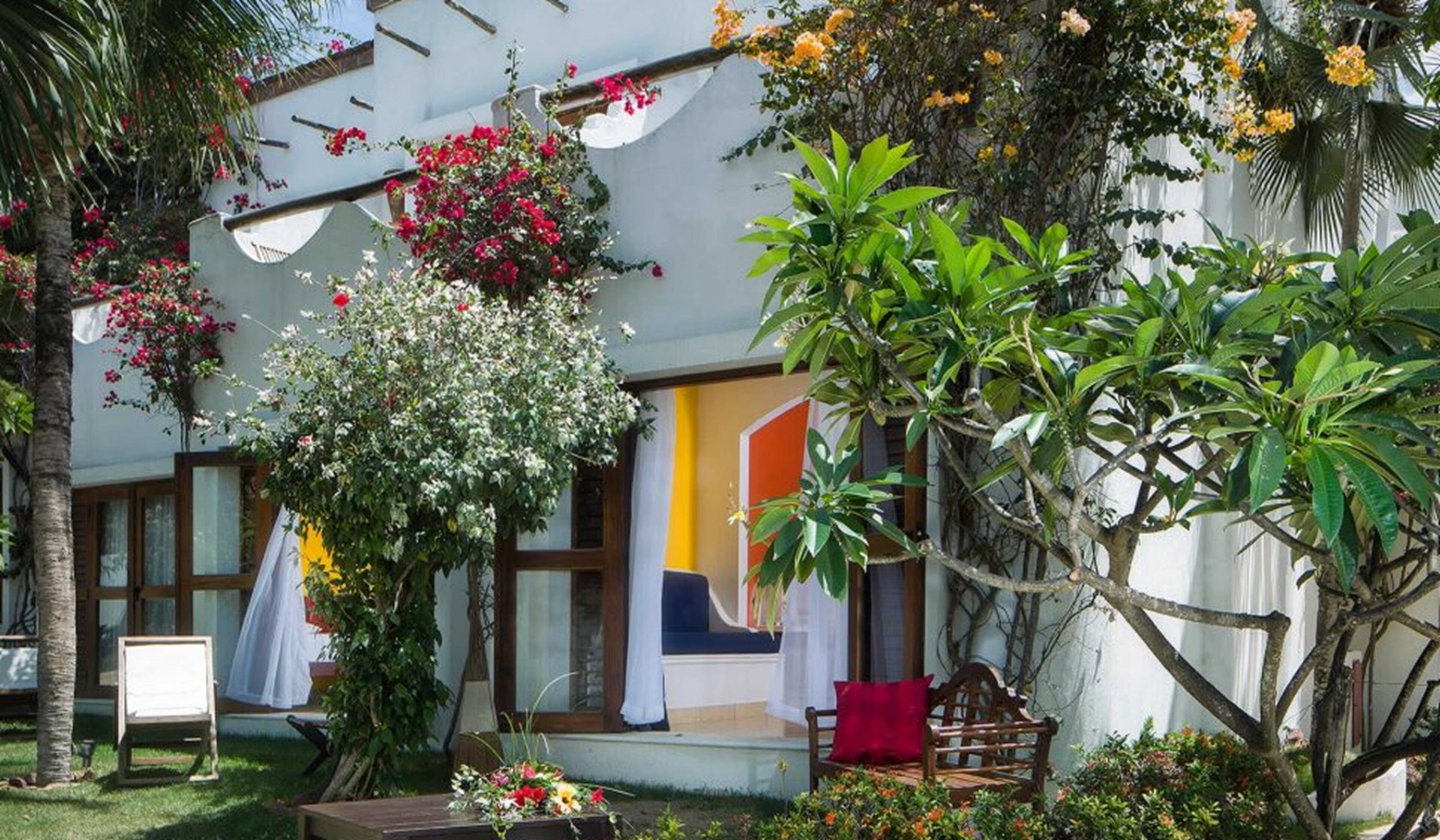 A cozy outdoor seating area with a bench, a table with flowers, and a view of a room with a blue couch and a yellow accent wall.