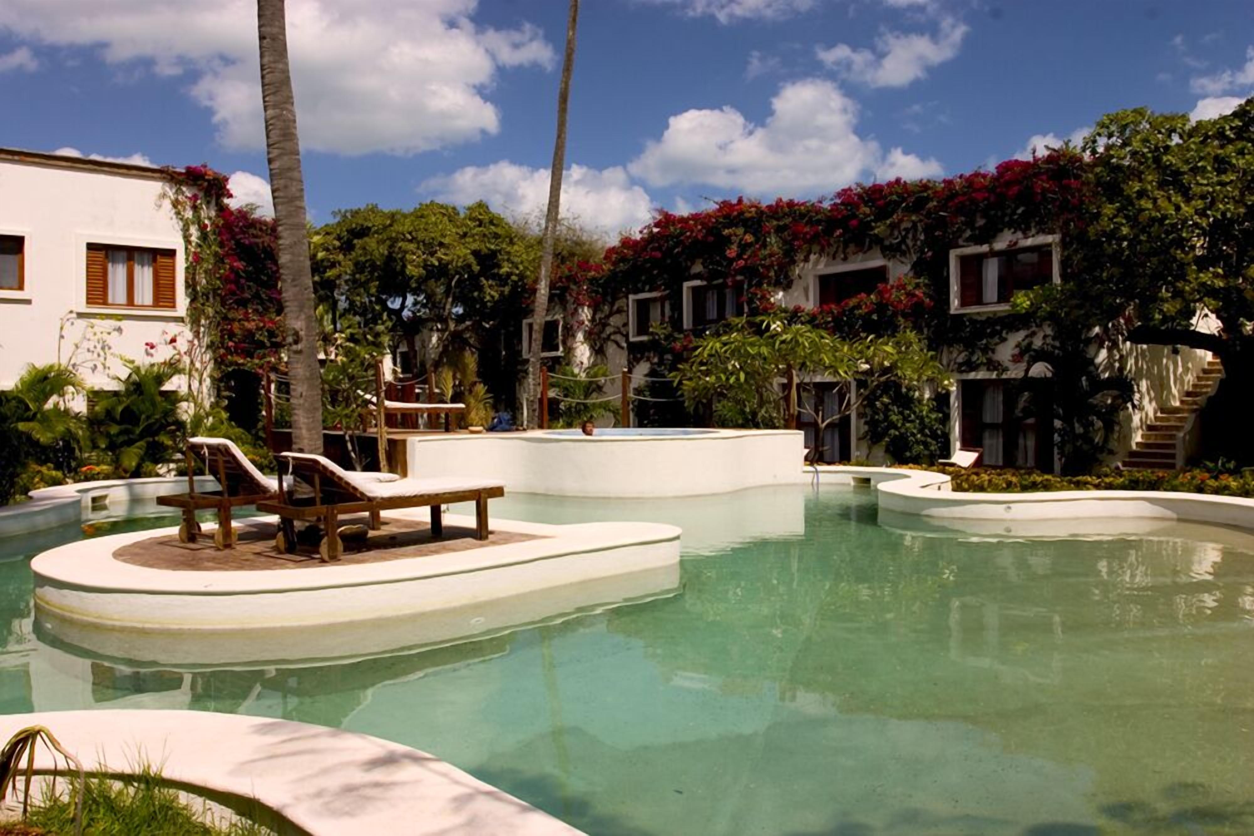 A pool area with lounge chairs and a bench, surrounded by buildings with red flowering vines.