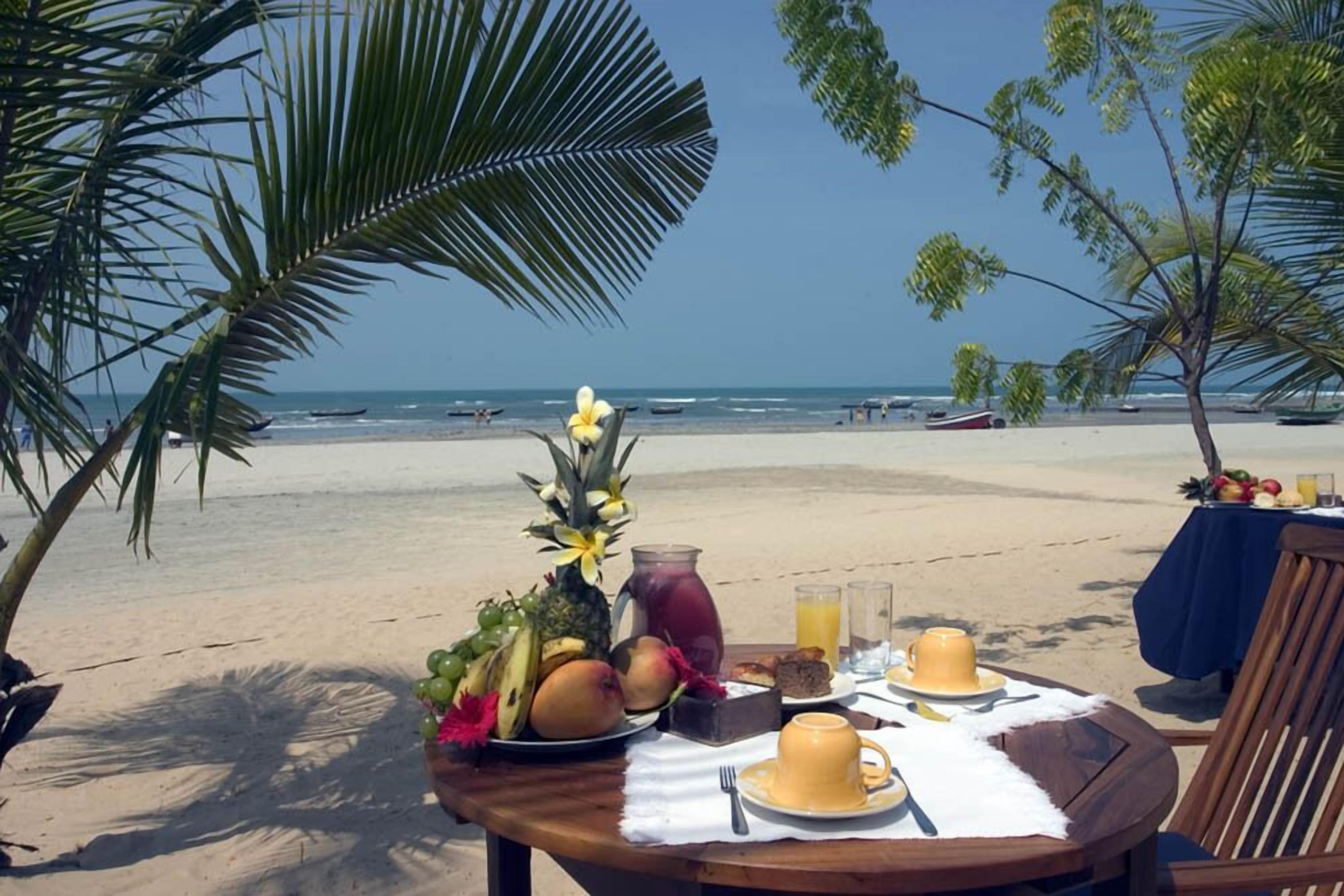 Una mesa frente al mar con alimentos para el desayuno, incluyendo frutas, una bebida y un plato de comida, con vista al océano.