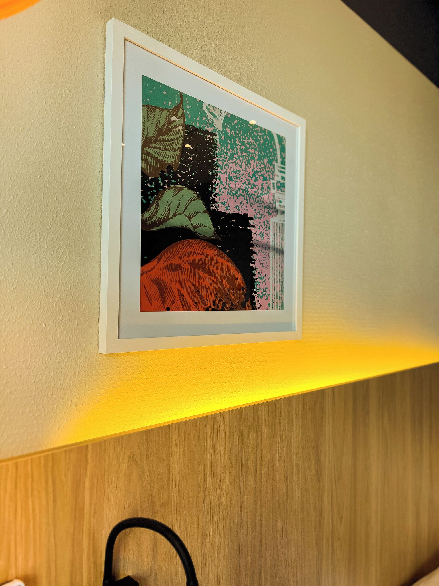 Een lijstwerk met een kunstwerk aan een muur, een houten paneel eronder, en een zwart object op de vloer.