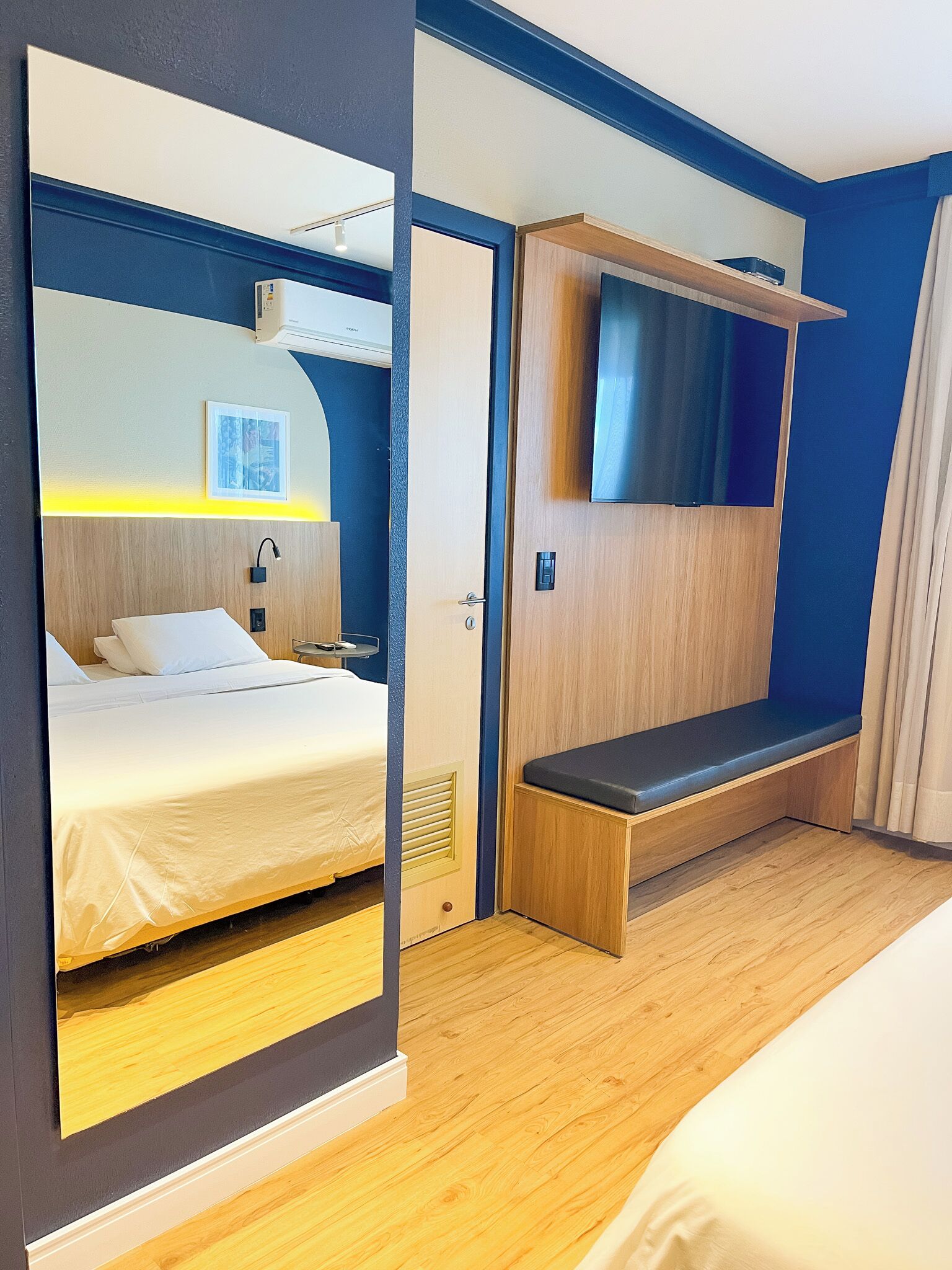 A hotel room with a bed, a bench, a TV, and a door.