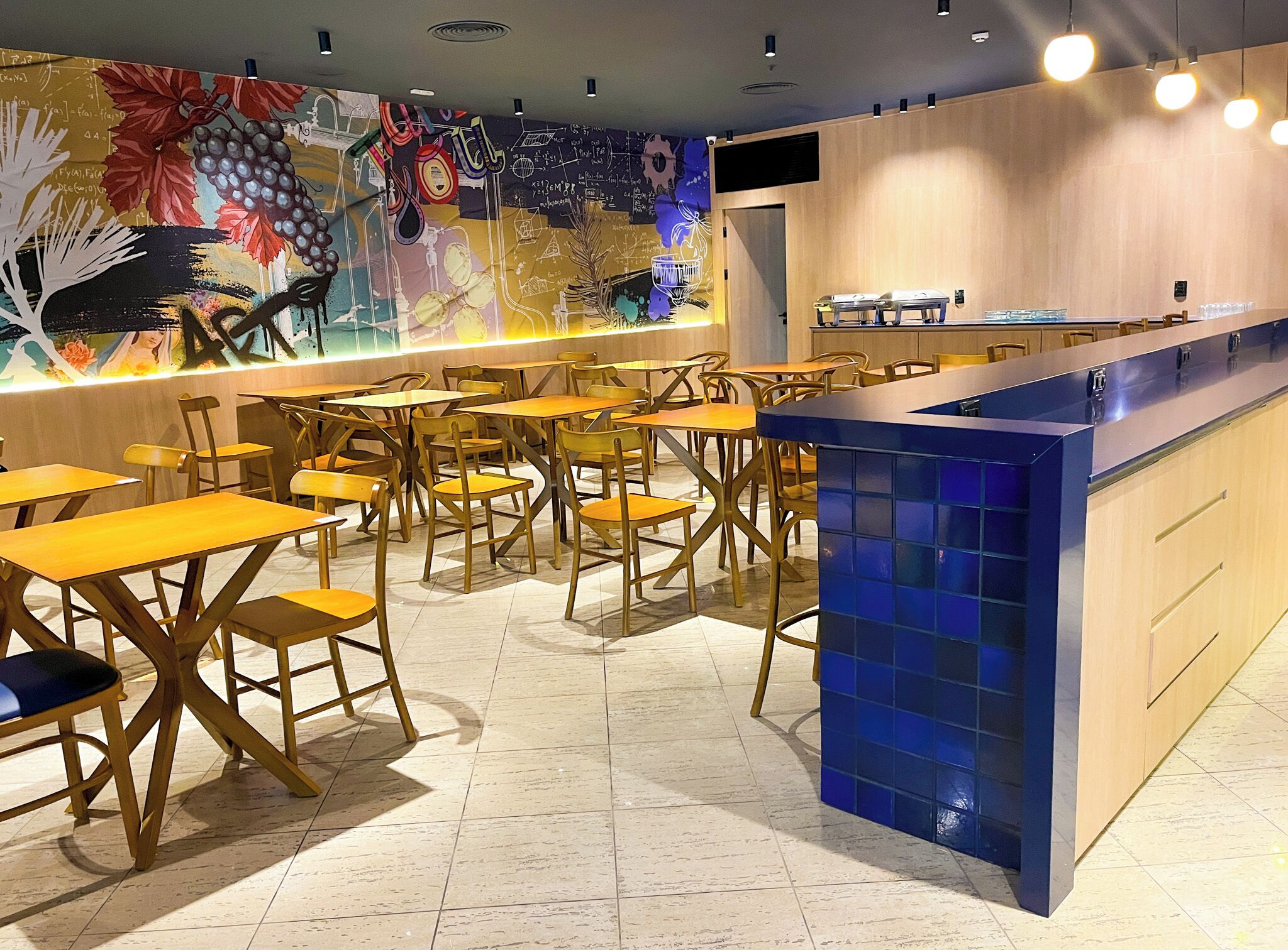 A restaurant interior with wooden tables and chairs, a blue-tiled counter, and a colorful mural on the wall.
