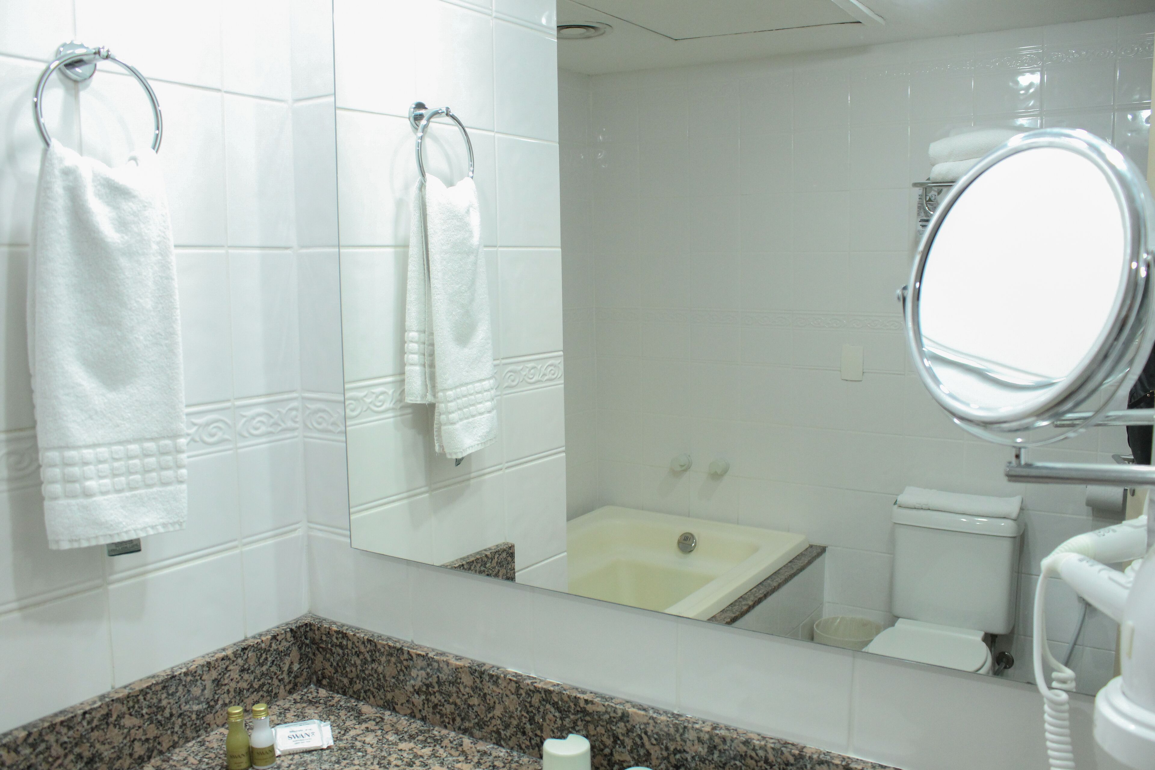 A bathroom with white tiles, a granite countertop, a sink, a mirror, a toilet, and a bathtub.