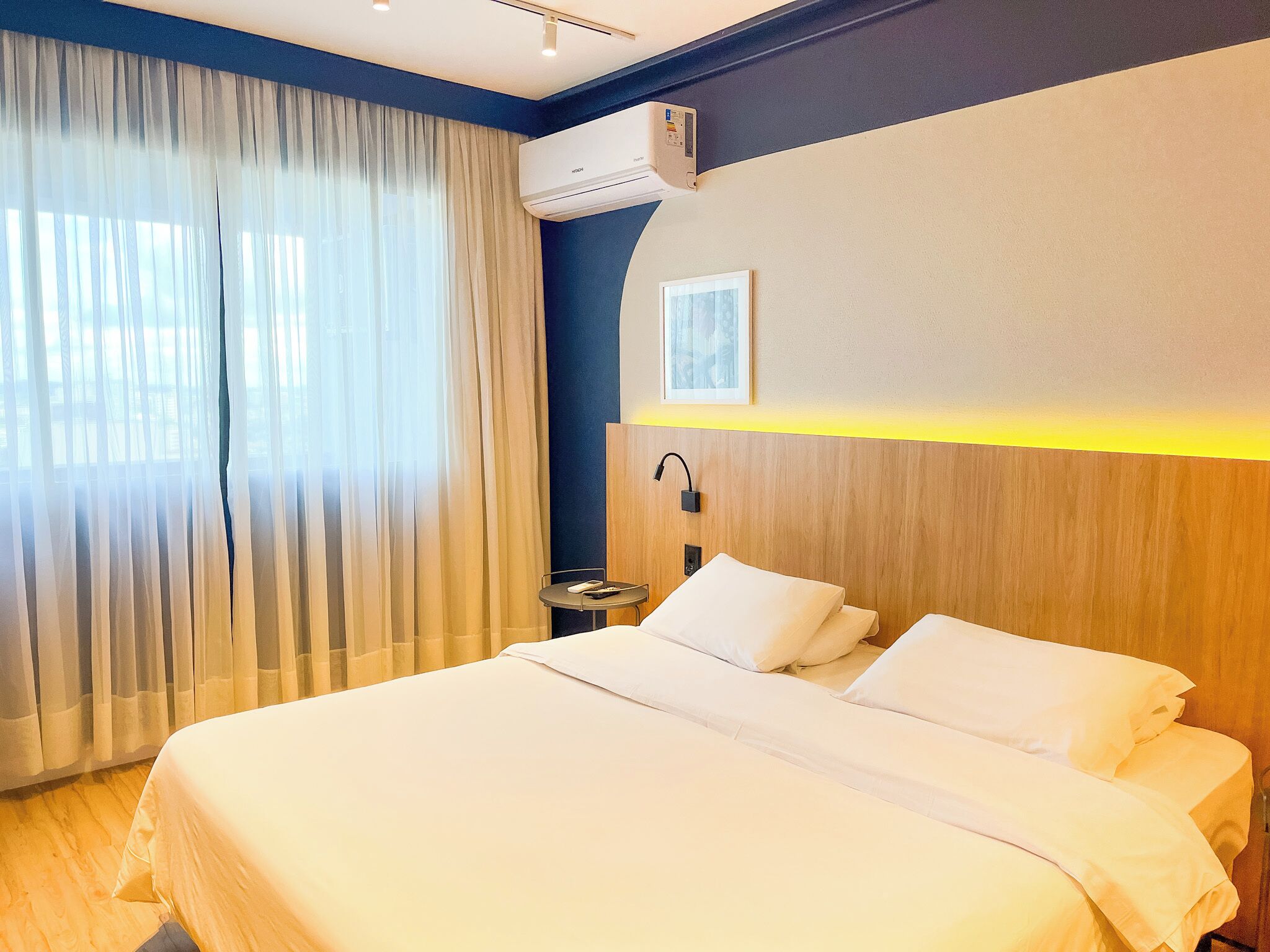A hotel room with a bed, curtains, an air conditioning unit, a bedside table, and a wall-mounted lamp.