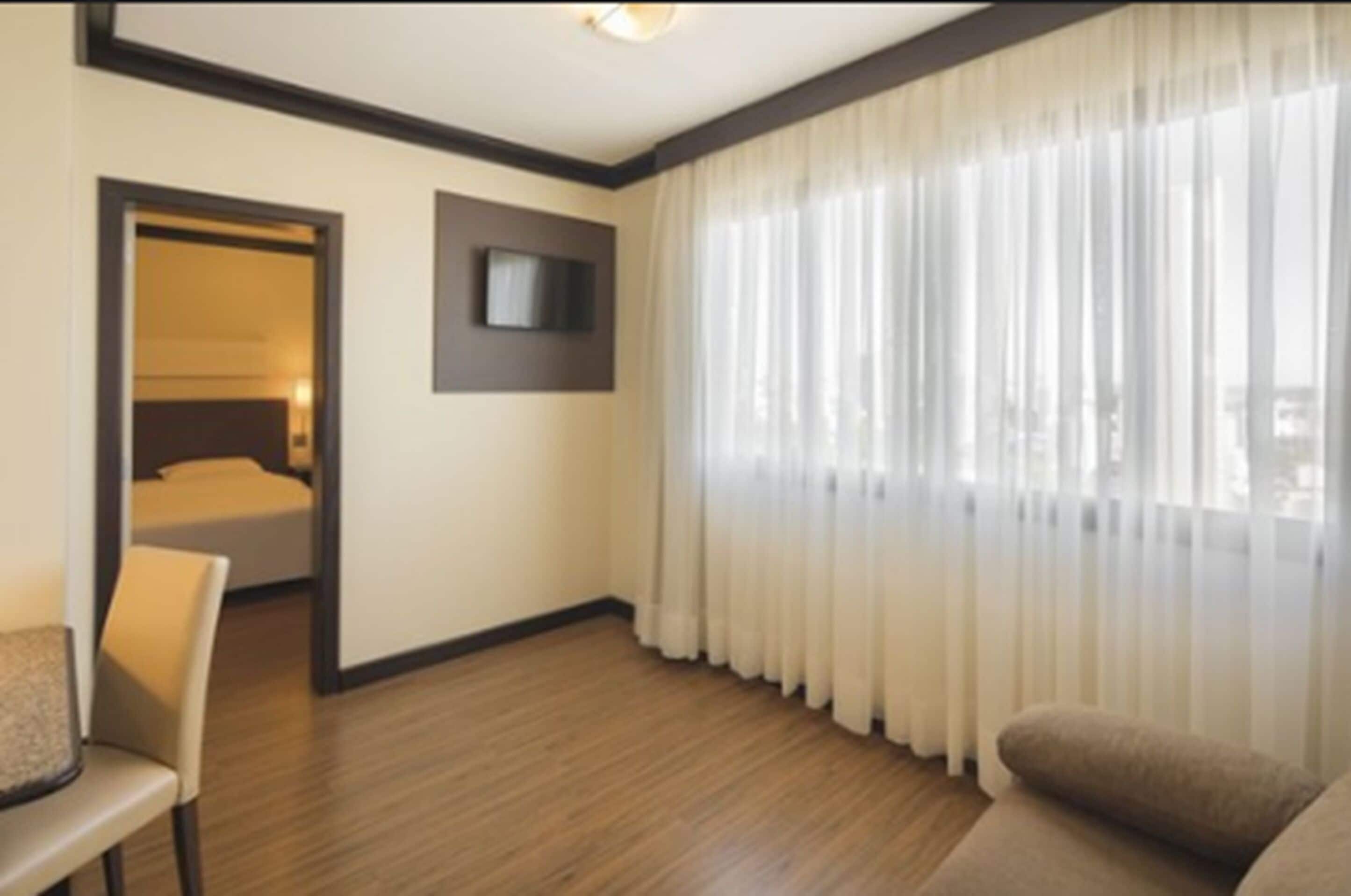 Ein Hotelzimmer mit Holzboden, einem Bett mit Kopfteil, einem an der Wand montierten Fernseher und einem großen Fenster mit transparenten Vorhängen.