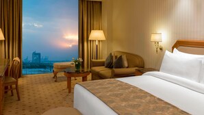 A hotel room with a large bed, a sofa, a desk, and a view of a cityscape at sunset. - Sheraton Kuwait, A Luxury Collection Hotel, Kuwait City (Kuwait City)