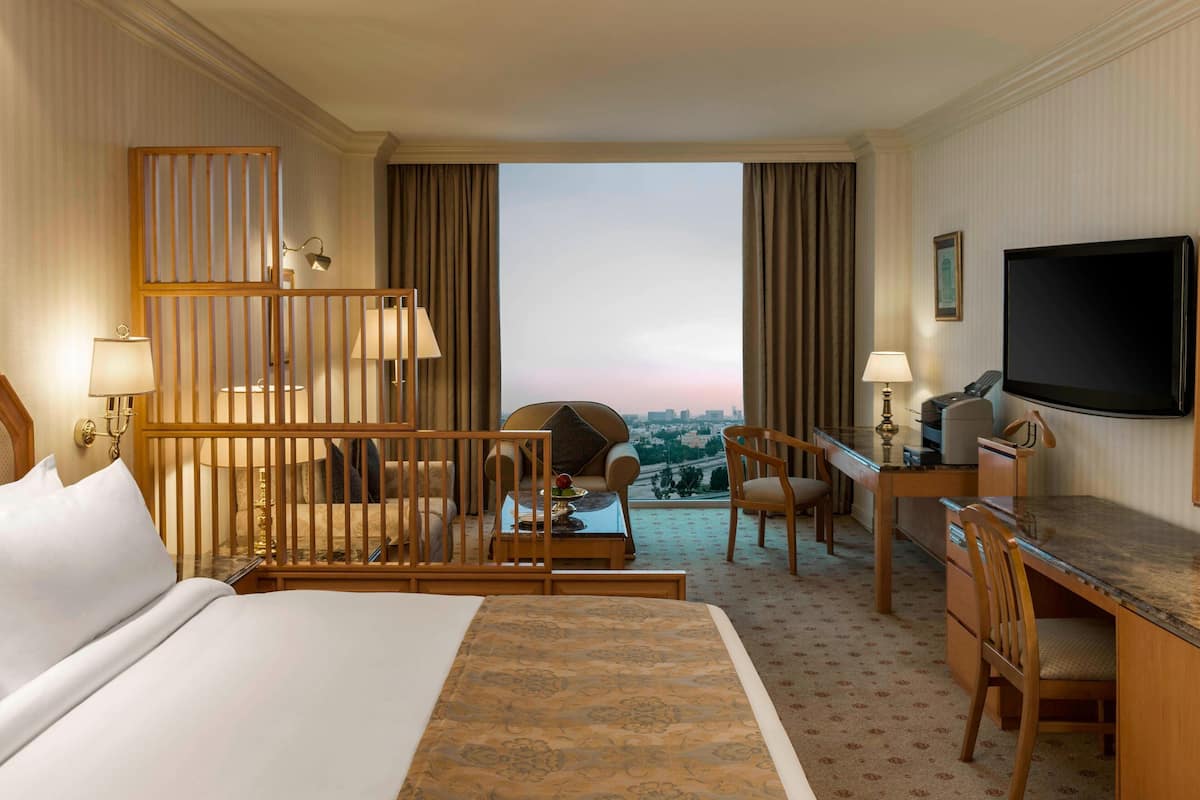 A hotel room with a large bed, a desk with a chair, a TV, and a view of the cityscape.