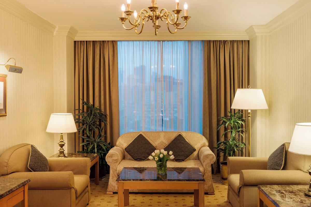 A hotel room with a sofa set, two lamps, a chandelier, and a view through sheer curtains.