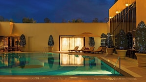 A swimming pool with lounge chairs and a covered seating area, surrounded by potted trees and a building with large windows. - Sheraton Kuwait, A Luxury Collection Hotel, Kuwait City (Kuwait City)