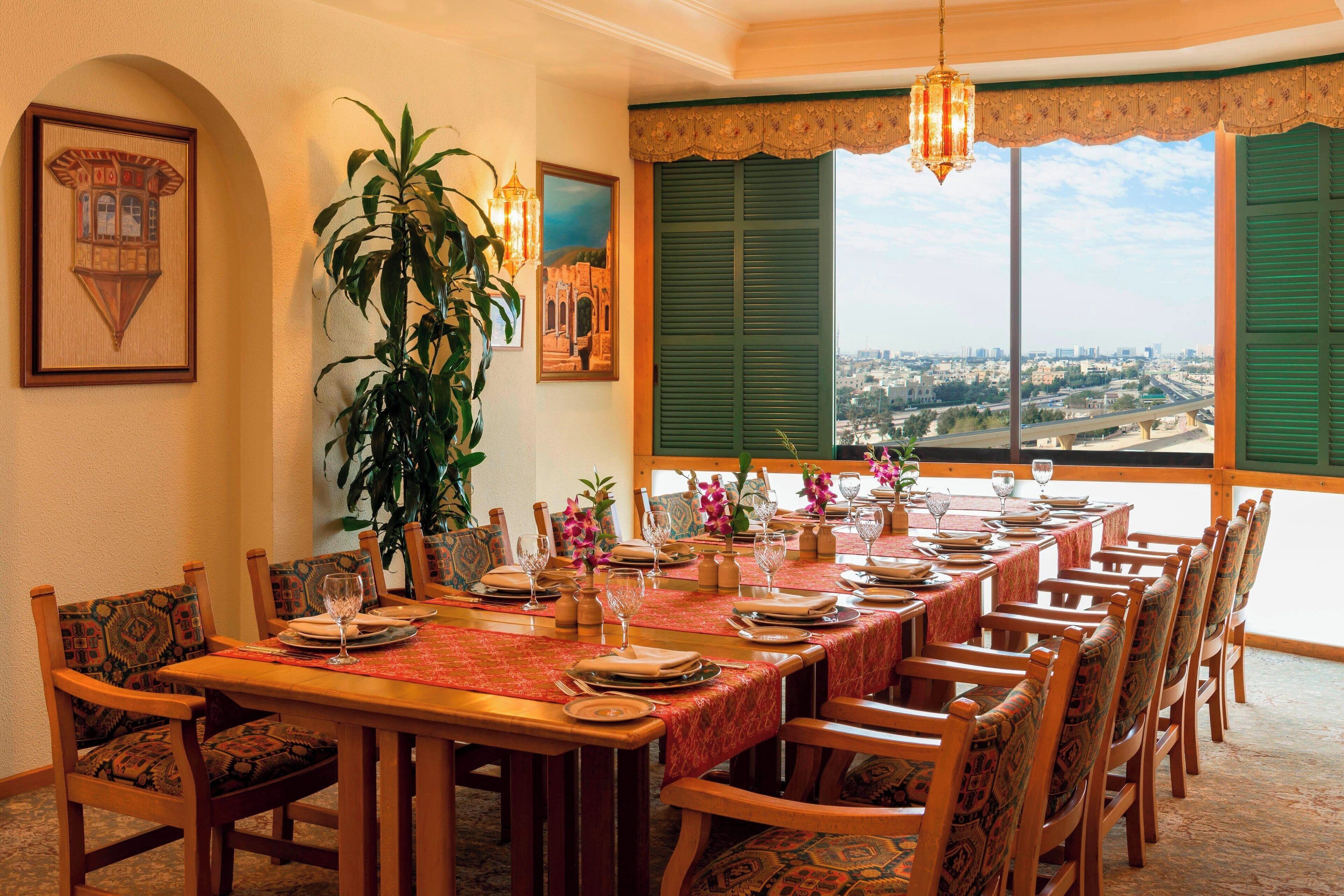 A dining area with a long table set for a meal, surrounded by chairs, with a city view through large windows.
