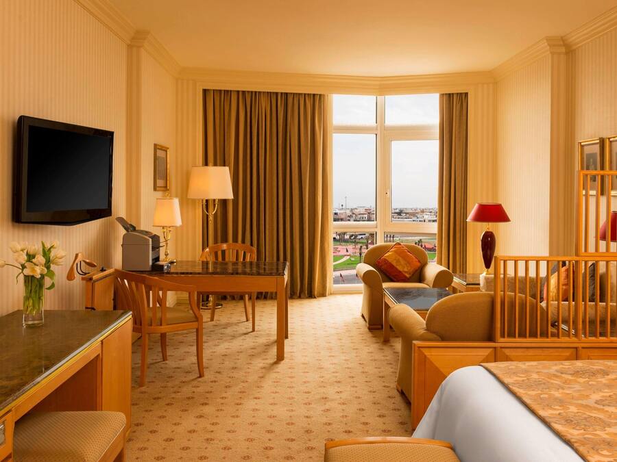 A hotel room with a bed, a desk, a chair, a table, a TV, a window with curtains, and a view of the city.