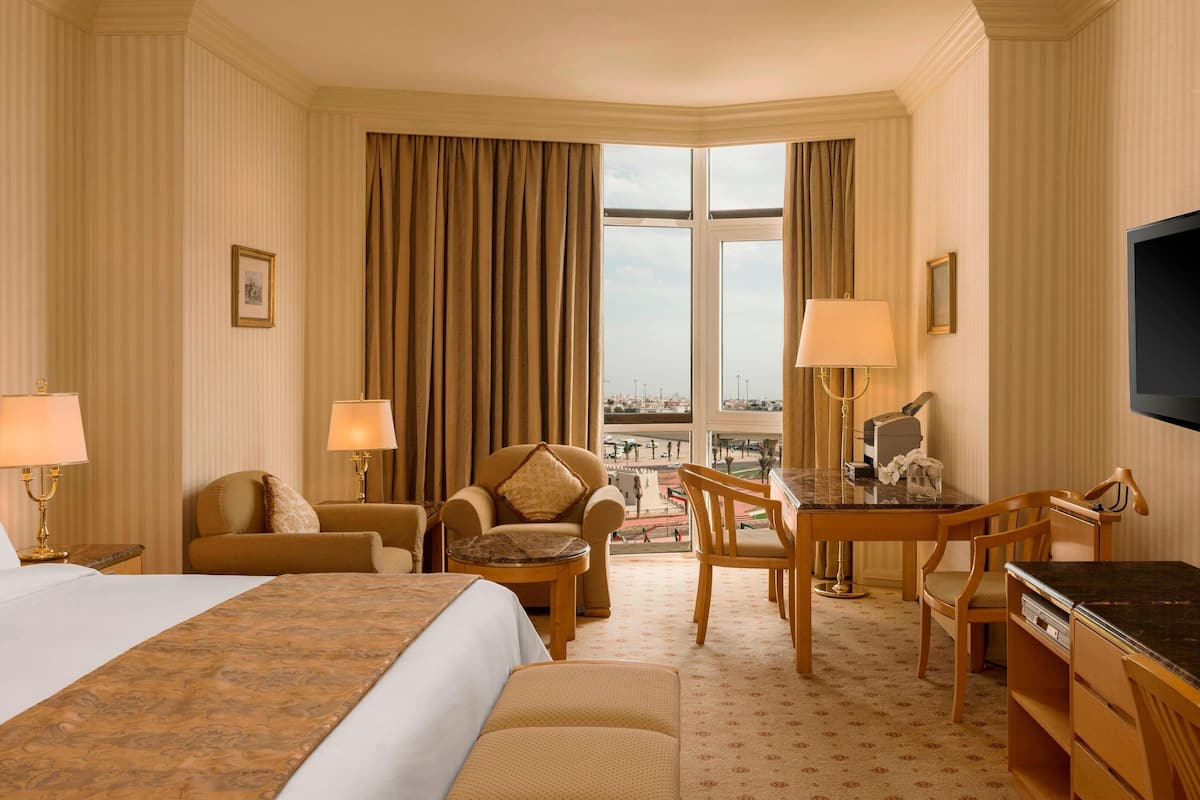 A hotel room with a large bed, two armchairs, a small table, a desk, and a TV. The room has a large window with curtains, a view of the cityscape, and a patterned carpet.