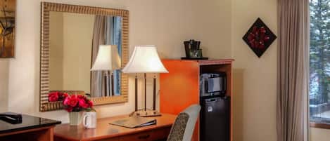A hotel room with a wooden desk, a chair, a small refrigerator, a microwave, a coffee maker, a vase with flowers, a mirror, and a window with curtains.
