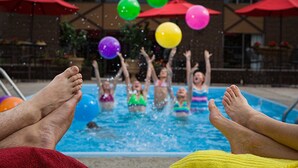 Vista de la piscina con una persona relajándose en una toalla amarilla, una piscina y sombrillas rojas al fondo.
