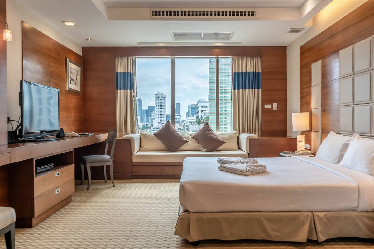 Habitación de hotel con cama, escritorio con computadora, silla, televisor y ventana grande con vista a la ciudad.