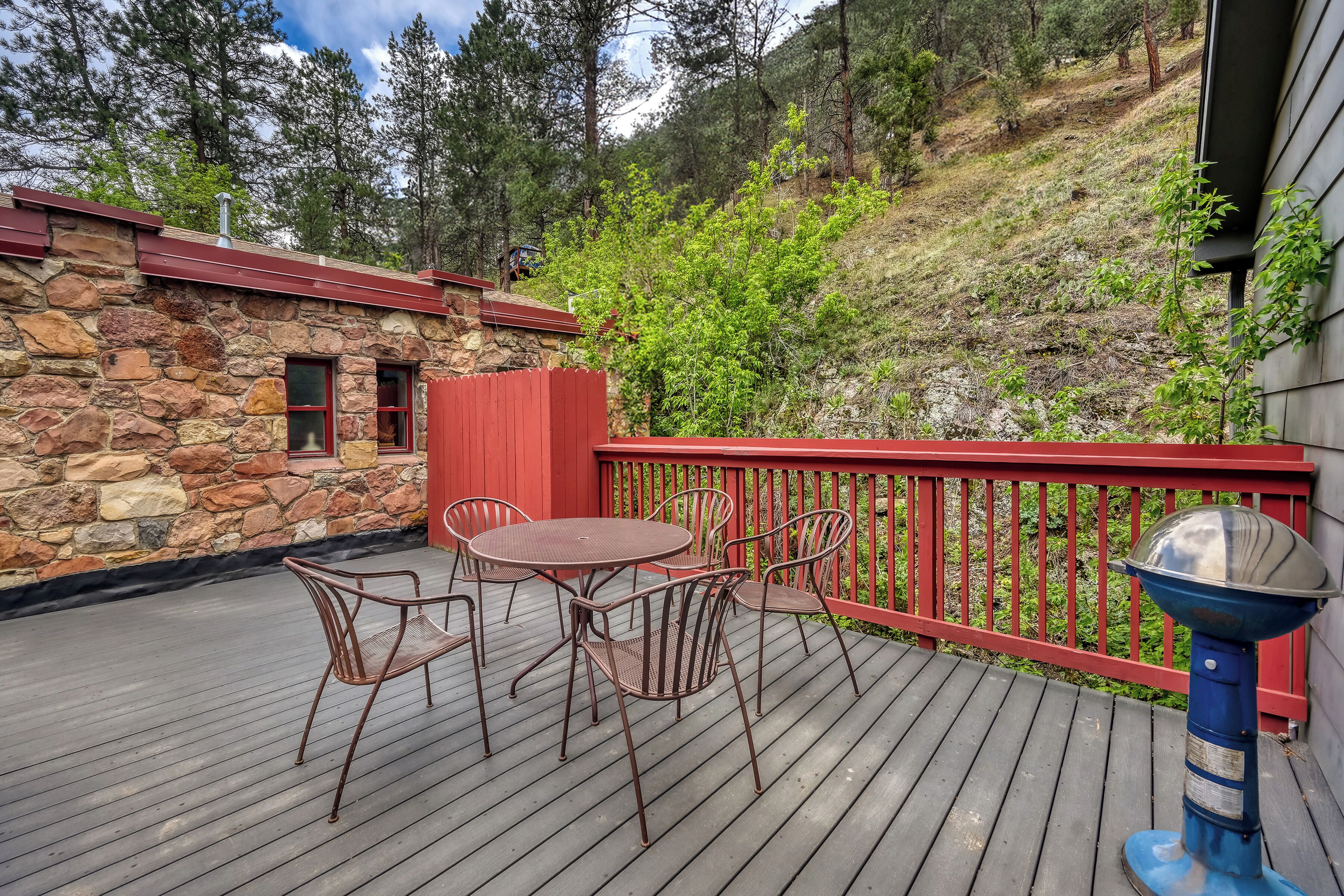 A wooden deck with metal chairs and a table, a stone building with a red roof, and a red railing.